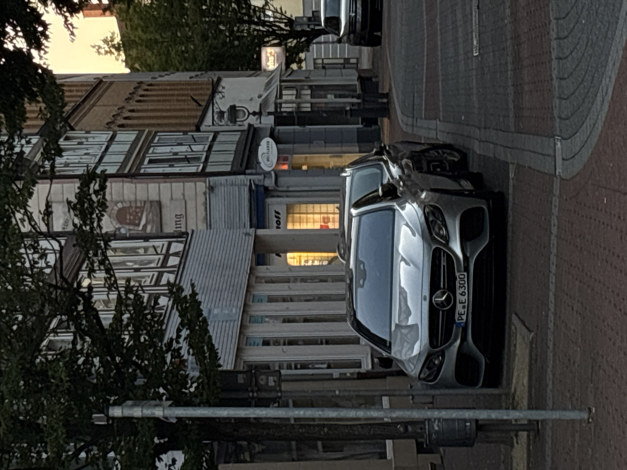 The image shows a street scene with parked cars and buildings. In the foreground, a silver Mercedes-Benz E-Class sedan with the license plate "PE E 6300" is parked on the right side of the street. The car has its headlights on. To the left of the Mercedes, another car is partially visible, a dark-colored SUV. The street is paved with cobblestones and has a curb.
In the background, there are several buildings. The building immediately behind the cars features a white facade with dark trim and windows. Further back, a taller building with a brick facade and arched windows is visible. Trees with dark green foliage are present in the scene, partially obscuring the buildings. The sky is overcast, with a hint of sunset colors towards the right side of the image. The overall lighting suggests it is late afternoon or early evening. There are no people visible in the image.