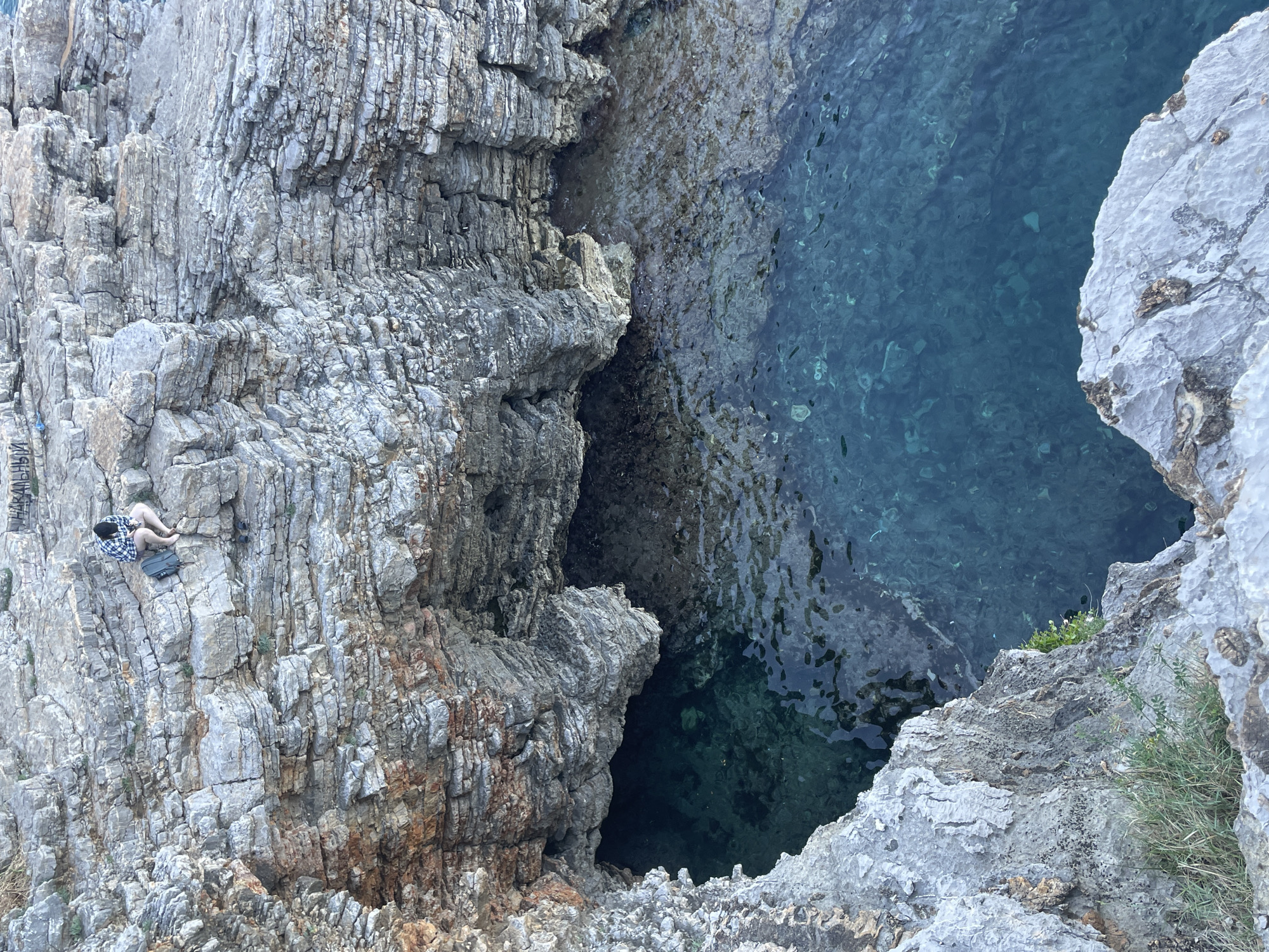 A high-angle shot presents a rugged coastal scene in Budva, Montenegro, focusing on a rocky outcrop meeting the Adriatic Sea. The left side features a layered rock formation where a person sits near the top edge of the frame. The individual is clad in dark garments, with a plaid shirt, a dark hat and a bag. Beside them, there is some writing on the rock face, obscured by the rock formations. The rocks show gray and brown hues with vertical striations. To the right, clear blue water fills a gap between the rocky edges. Some foliage grows on the rocks near the water. The overall lighting is bright, highlighting the texture of the rocks and clarity of the water.