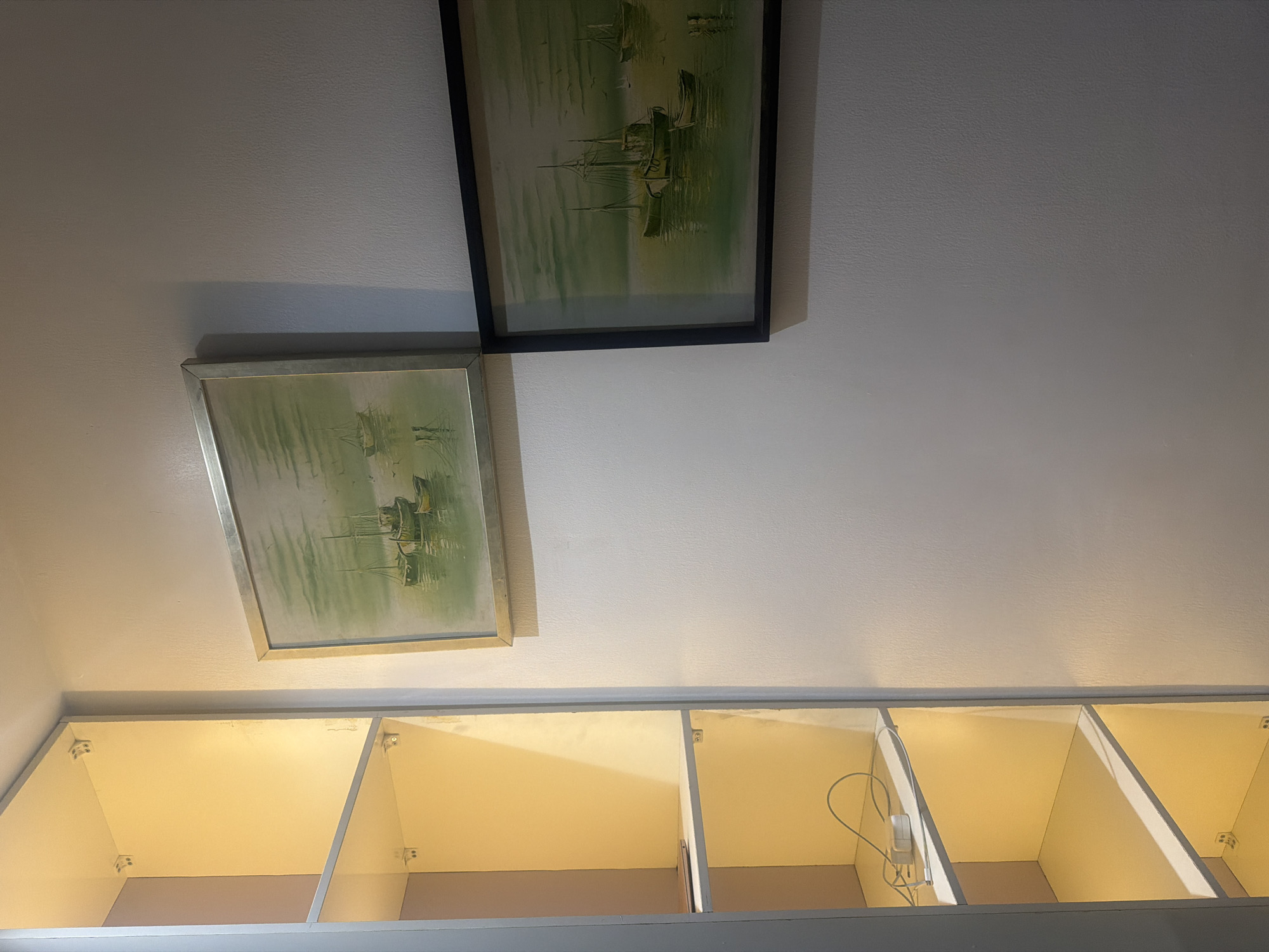 The image displays an indoor scene featuring two framed paintings and a row of illuminated cubby shelves. Two framed pictures hang on a light-colored, textured wall. The upper painting, partially visible, has a black frame and depicts a nautical scene with green-hued water and boats. Below and to the left, another painting with a lighter, possibly silver, frame shows a similar scene of boats reflected on calm, green water.
Below the paintings, a long, horizontal structure contains multiple open cubby compartments. These cubbies are uniformly lit from within by a warm, yellow light, indicating internal lighting fixtures. Inside one of the rightmost cubbies, a white electronic device, possibly a charger or small light, with a cable is visible. The cubbies appear to be part of an overhead cabinet or shelving unit. No people are present, and there is no visible text. The scene is static, showcasing interior decor.