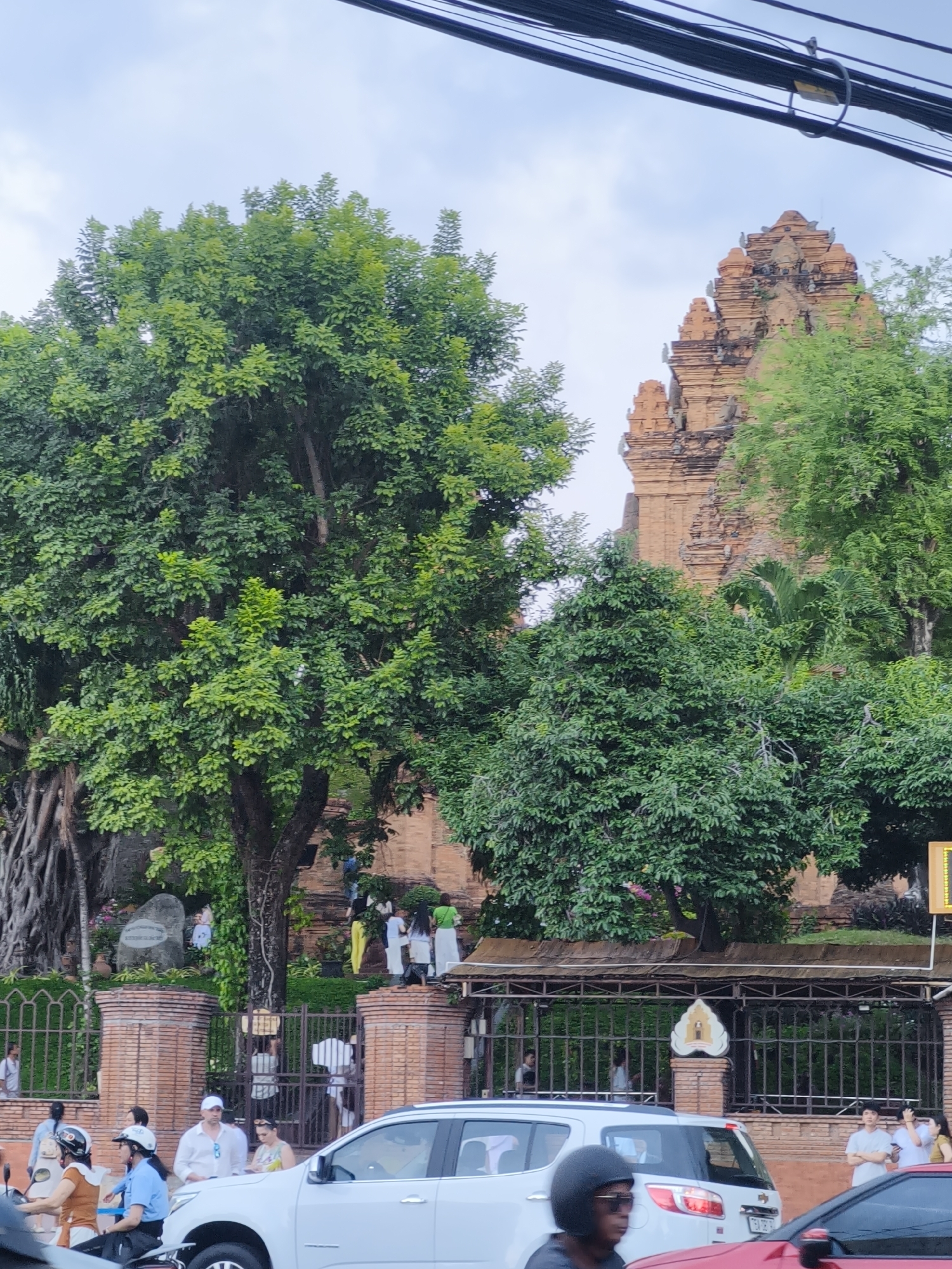 This media file captures a scene in Bac Nha Trang Ward, Vietnam, featuring a view of the Po Nagar Cham Towers, a historical landmark constructed from reddish-brown brick. The towers rise majestically against a cloudy sky. In the foreground, a lush green tree with thick foliage dominates the left side of the frame, its exposed roots visible near the base. Another mature tree stands to the right, its branches partially obscuring the towers.
A group of people are seen walking up stairs towards the towers, suggesting they are visitors exploring the site. Some individuals are also visible on a pathway near the entrance, which is bordered by a brick wall and a metal fence. Cars, including a white SUV and a red vehicle, are present on a road in the lower portion of the image, indicating this is a public area accessible by vehicles. Several people are either driving or waiting near their vehicles.
The scene appears to be set during the daytime, with overcast conditions suggested by the bright but diffused light. The overall atmosphere is that of a typical day at a popular tourist or historical site, with people engaging in sightseeing and commuting. No specific text is clearly discernible in the image.