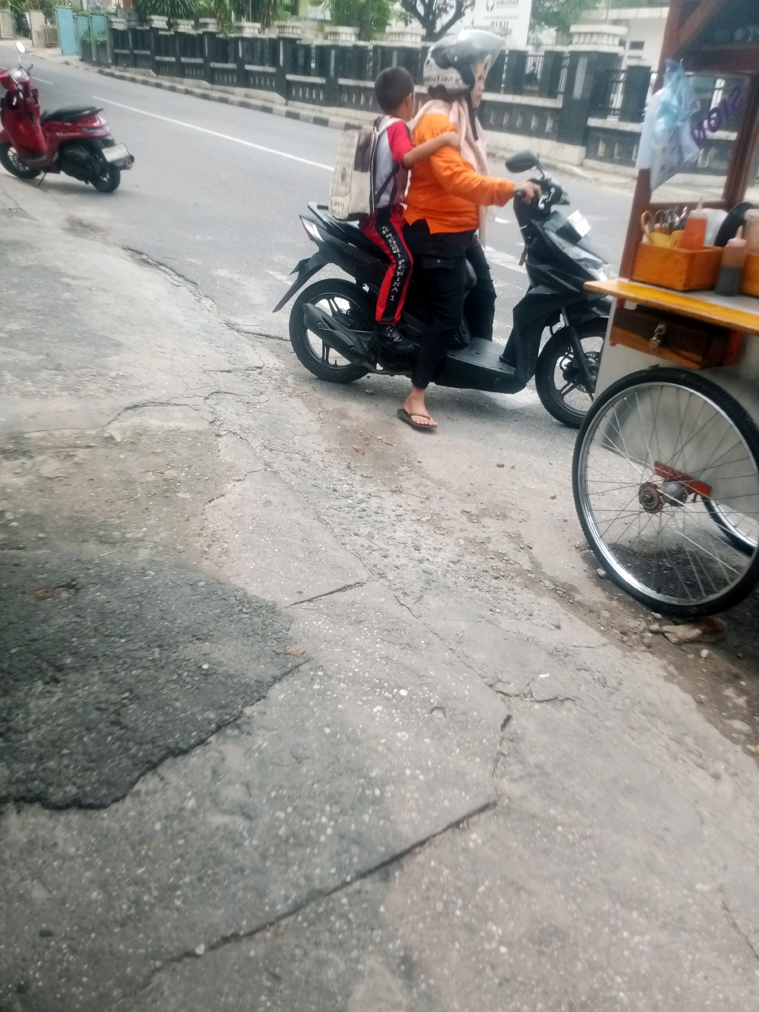 Two people are on a black motorcycle. The person in front is wearing an orange shirt and black pants, with a light pink scarf around their neck. They are wearing a silver helmet. The person behind them is wearing a red shirt and black pants with a white stripe. They have a backpack with white and brown straps. They are riding on a paved street. To the right of the motorcycle is a food cart with a wooden frame. The street has some cracks and damage. A red scooter is parked on the left side of the street. The background shows a fence and some greenery, suggesting an urban or suburban environment. The image appears to be taken in Pekanbaru, Indonesia, based on the style of the motorcycles and the presence of a food cart commonly found in that region.