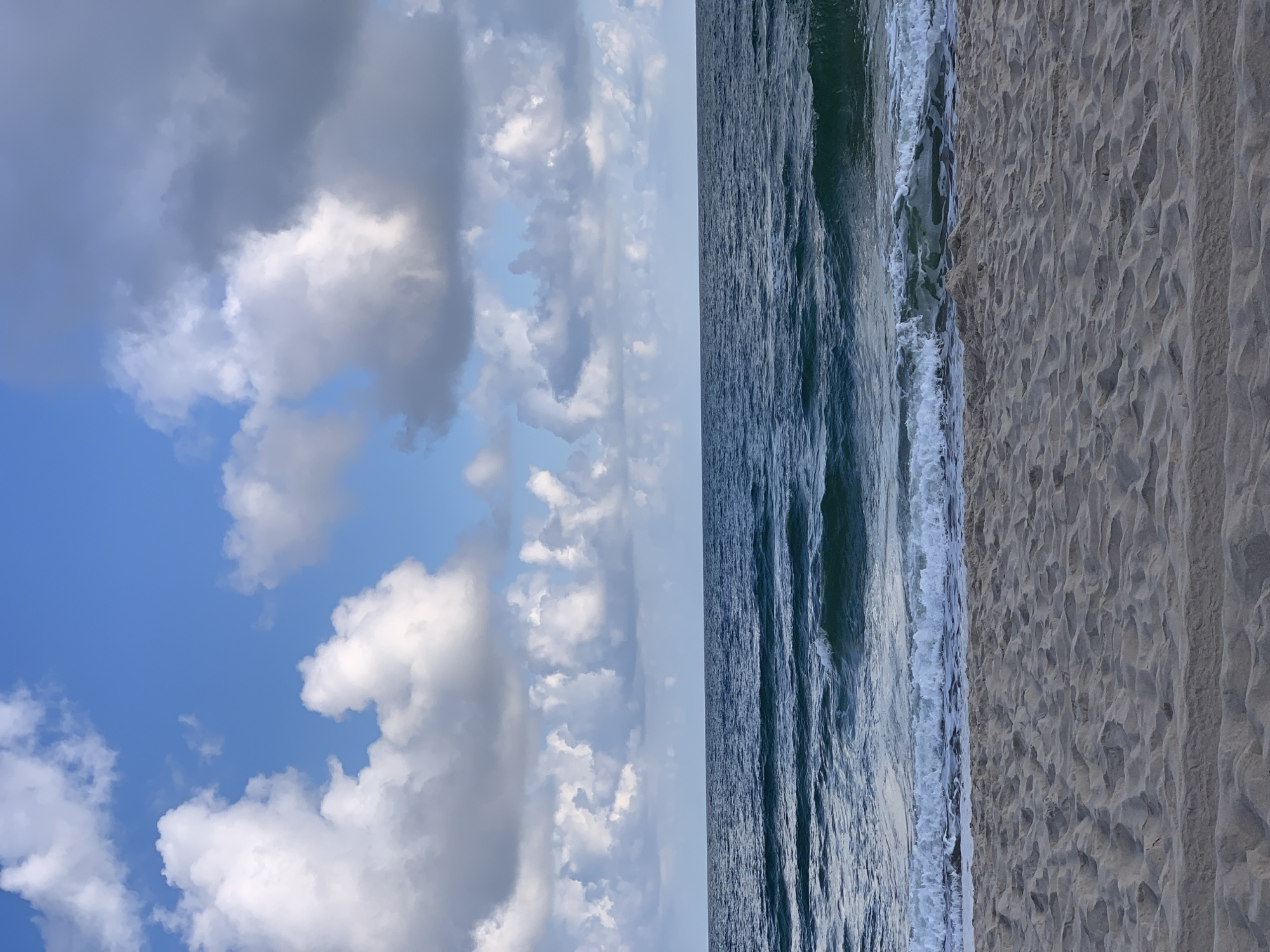 This is an image of a beach scene. The foreground consists of a sandy beach with visible footprints and ripples in the sand. The middle ground is occupied by the ocean, with waves gently breaking on the shore. The water is a deep blue-green color. The background is a sky filled with a mix of white and gray clouds against a clear blue sky. The scene depicts a calm day at the coast. The location is not specified in the image, therefore it cannot be identified as Rewal, Poland.