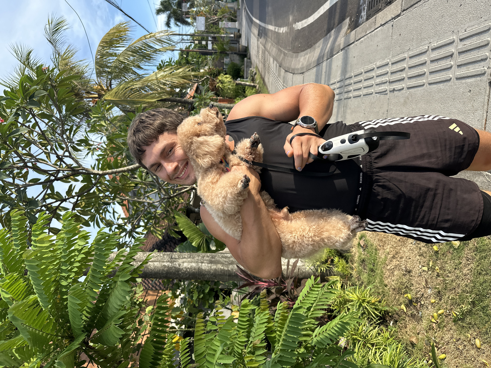A medium shot depicts a smiling, light-skinned male in an outdoor setting that appears to be located in North Kuta, Indonesia. The man is holding a tan-colored poodle and holding a white and black object that has a handle. The man is wearing a black tank top and black shorts with white stripes. He also has a watch on his left wrist. The environment includes trees, lush green plants, a paved road, and buildings. The man and dog are positioned on the side of the road, near dense vegetation.