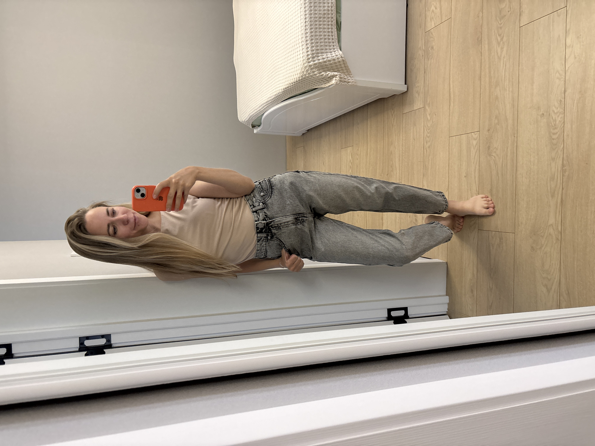 A young Caucasian woman with long blonde hair is standing in a room, taking a mirror selfie. She is wearing a light beige sleeveless top and distressed grey denim jeans. Her right hand is holding an orange-cased smartphone. The room has light wooden flooring and pale grey walls. To the right, a white bed with a light-colored waffle-weave blanket is visible. To the left, a white door frame with black hinges is present. The image appears to be taken indoors, likely in a bedroom or dressing area. There is no indication that this location is in the city of Chaikovsky, Russia.