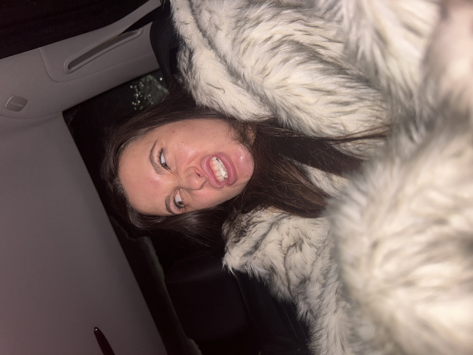 A single female individual is depicted inside a vehicle at night in Высокиничи, Russia. She has long, dark hair and is wearing a voluminous, light-colored faux fur coat with white and gray tones. The subject is making an exaggerated facial expression, characterized by wide, upward-angled eyes, a wrinkled nose, and an open mouth revealing clenched upper and lower front teeth in a grimace. Her gaze is directed towards the upper left of the frame. The background reveals the dark interior of the vehicle, including a headliner, a portion of an A-pillar with what appears to be an airbag indicator, and a window displaying blurred exterior lights and visible raindrops, suggesting precipitation outside.
