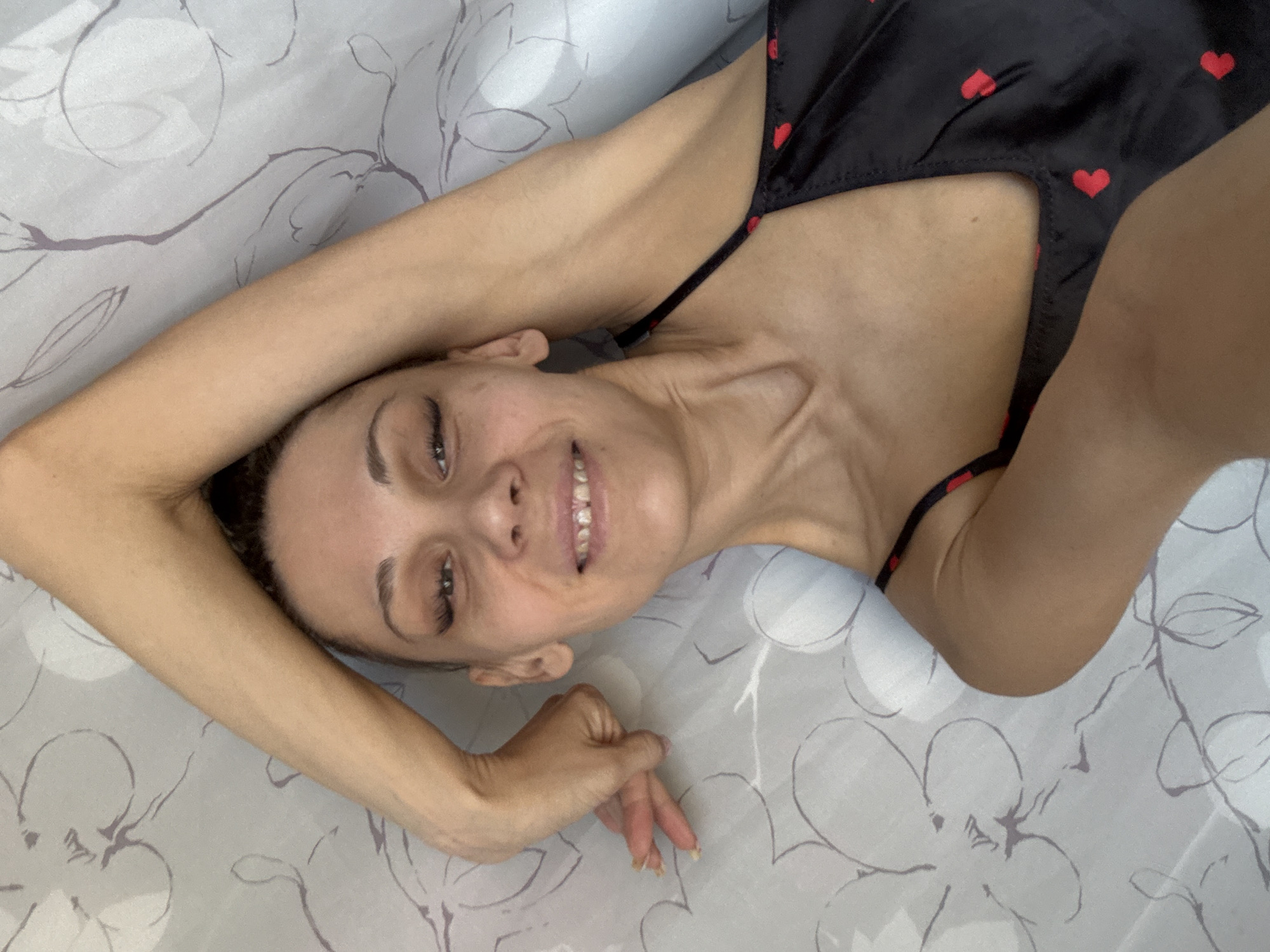 A fair-skinned woman with brown hair pulled back is lying on a light-colored bed with a floral pattern. She is wearing a black spaghetti strap top with small red hearts. She is smiling and looking directly at the camera. Her right arm is bent and raised above her head, with her hand near her face. Her left arm is bent at her side. The location is not identifiable from the image.