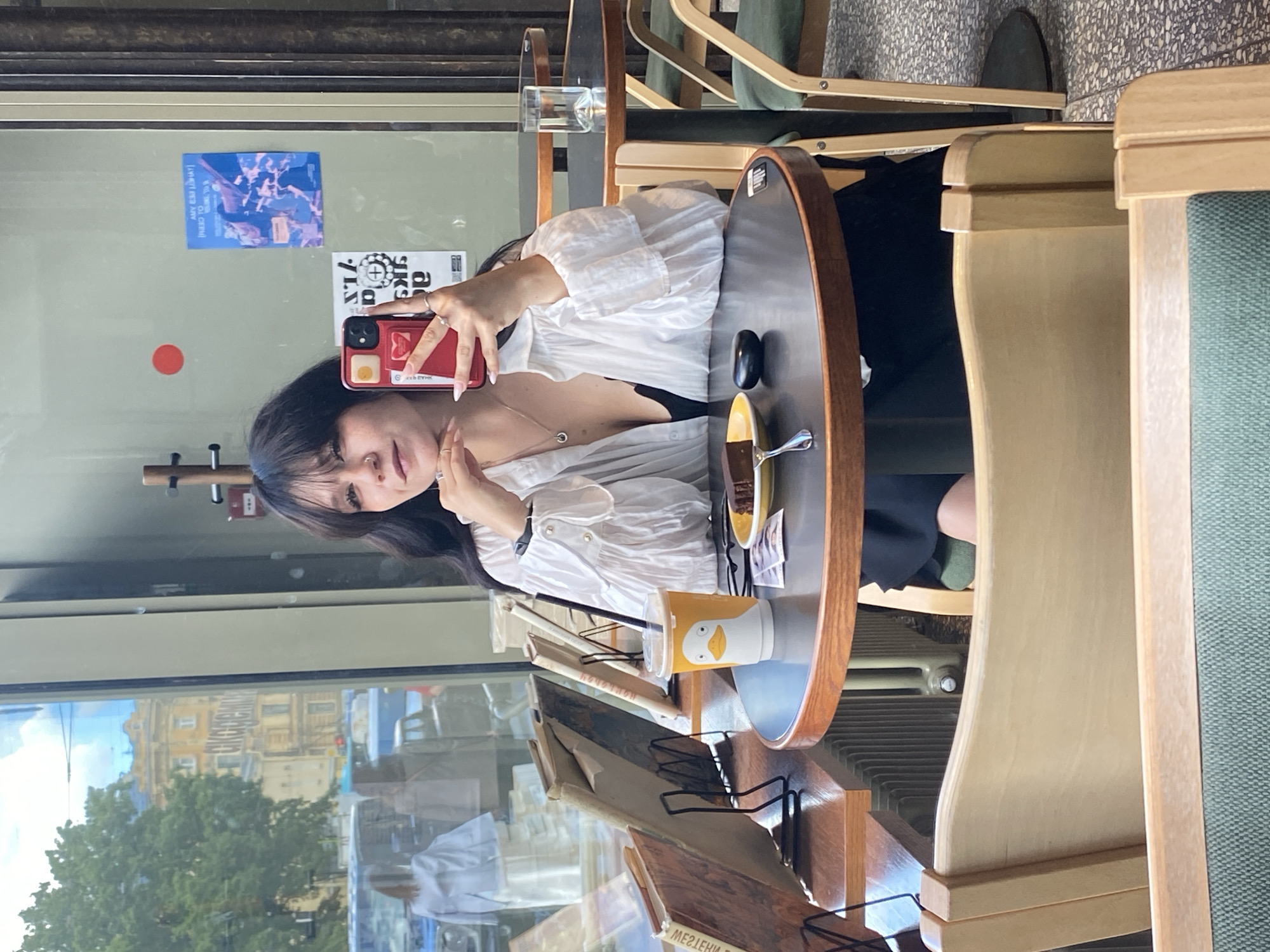 A young woman with long dark hair is taking a mirror selfie in a cafe with large windows. She is wearing a white blouse and a black top, and she is holding a red smartphone. On the table in front of her is a cup with a duck on it, a slice of chocolate cake on a yellow plate with a fork, and a small black object. Behind her, the cafe is visible, with other tables and chairs, and a display of books on a wooden shelf. Through the window, a street scene in Saint Petersburg, Russia, is visible, with buildings and trees.