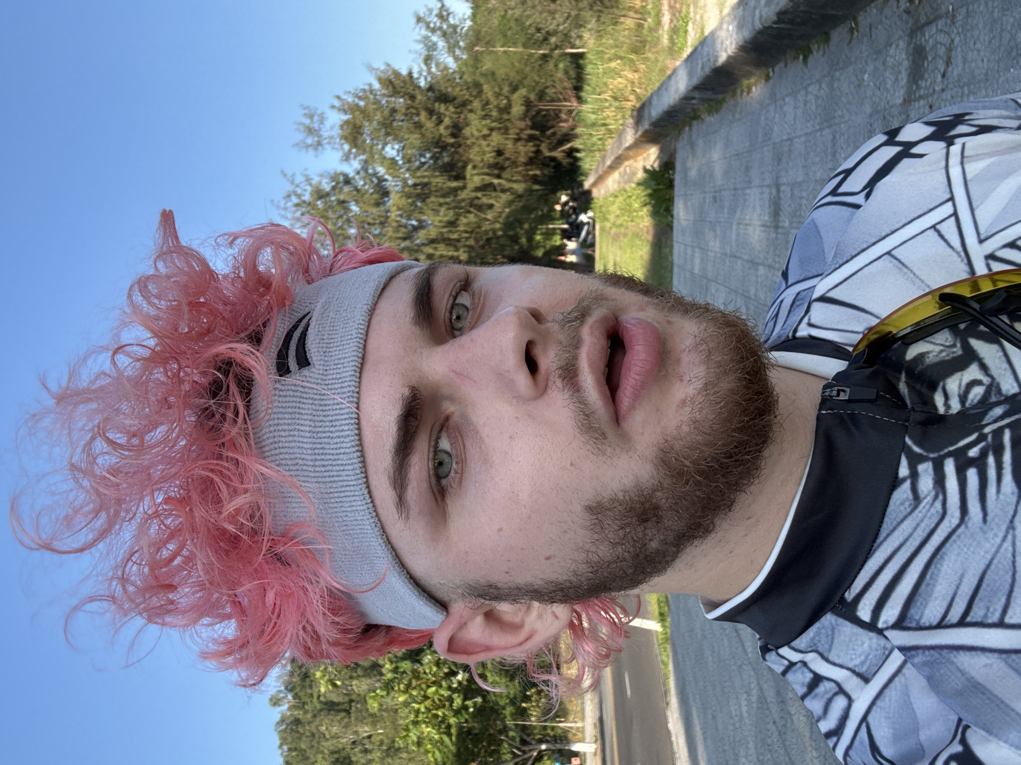 A selfie of a young man with pink hair, light eyes, and a beard wearing a grey headband. He is wearing a black and white patterned shirt. Behind him is a paved path, a grassy area, trees, and a blue sky. There is nothing in the image that indicates it is located in the city of Da Nang, Vietnam.