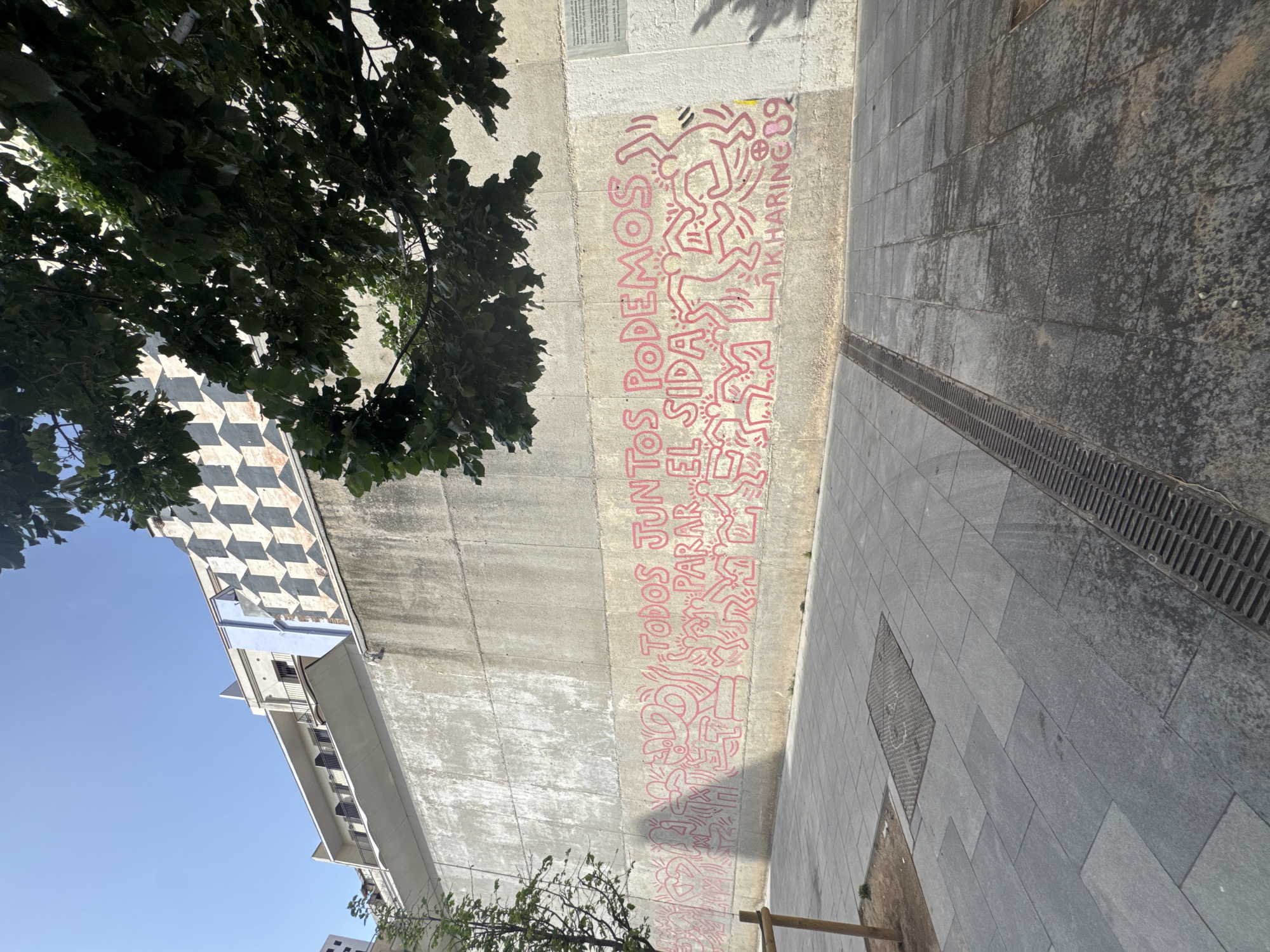 The image displays an urban outdoor scene featuring a large concrete wall adorned with a mural by artist Keith Haring. The mural is rendered in bright red/pink lines against the grey concrete, depicting numerous iconic dancing and interacting stick figures in dynamic poses. Prominently displayed text in Spanish reads "TODOS JUNTOS PODEMOS PARAR EL SIDA" (All Together We Can Stop AIDS), with the artist's signature and year "K.HARING 89" visible in the upper right section of the mural.
To the left of the wall, a paved area is visible, composed of dark grey rectangular and square tiles, along with a long metal drainage grate. Overhead, lush green tree foliage densely covers the upper right portion of the image, casting shadows on the wall and ground. Through the gaps in the leaves, a building with a distinctive black and white checkered pattern on its upper facade is partially visible against a clear blue sky.
The scene is well-lit by bright sunlight, indicating a daytime setting and clear weather. This specific public artwork by Keith Haring is a significant piece dedicated to AIDS awareness and is located in Barcelona, Spain.