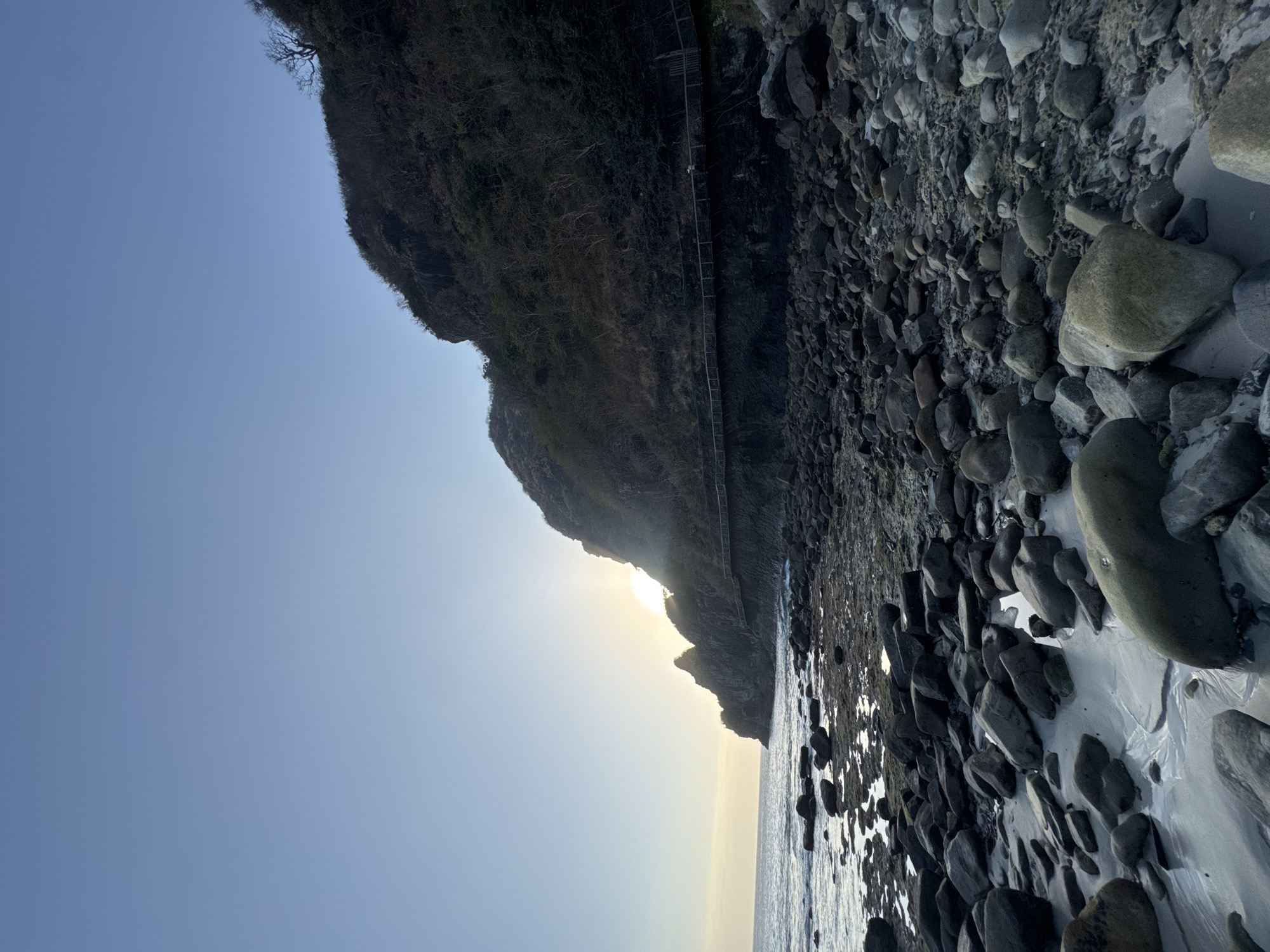 A vertical, low-angle shot captures a rocky shoreline at sunset in Sekongkang, Indonesia. The sun is low in the sky, peeking out from behind a dark, mountainous cliff face. A wooden boardwalk is visible traversing the cliff face, with a lone figure walking on it. The foreground is filled with smooth, gray rocks of various sizes scattered across the sand. The ocean stretches out to the left, with gentle waves lapping at the shore. The sky is a clear, pale blue, transitioning to a warmer yellow hue near the horizon. The overall impression is one of natural beauty and tranquility.