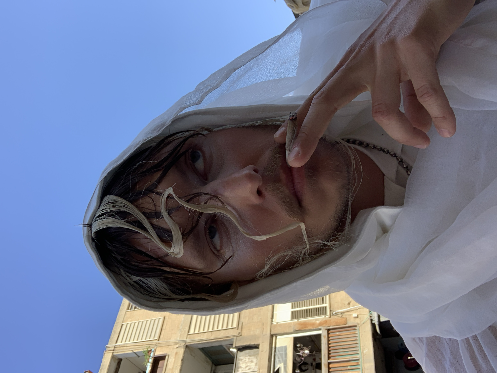 A low-angle medium shot depicts a person smoking a joint while standing on a balcony in Tel-Aviv, Israel. The person, likely male, has dark hair with blonde streaks, light skin, and stubble. They are wearing a white hooded robe. Their right hand is raised, holding a lit joint between the index and middle fingers. A silver chain is visible around their neck. Behind the person, a blue sky contrasts with the tan-colored apartment building, complete with visible windows, shutters, and electrical wiring.