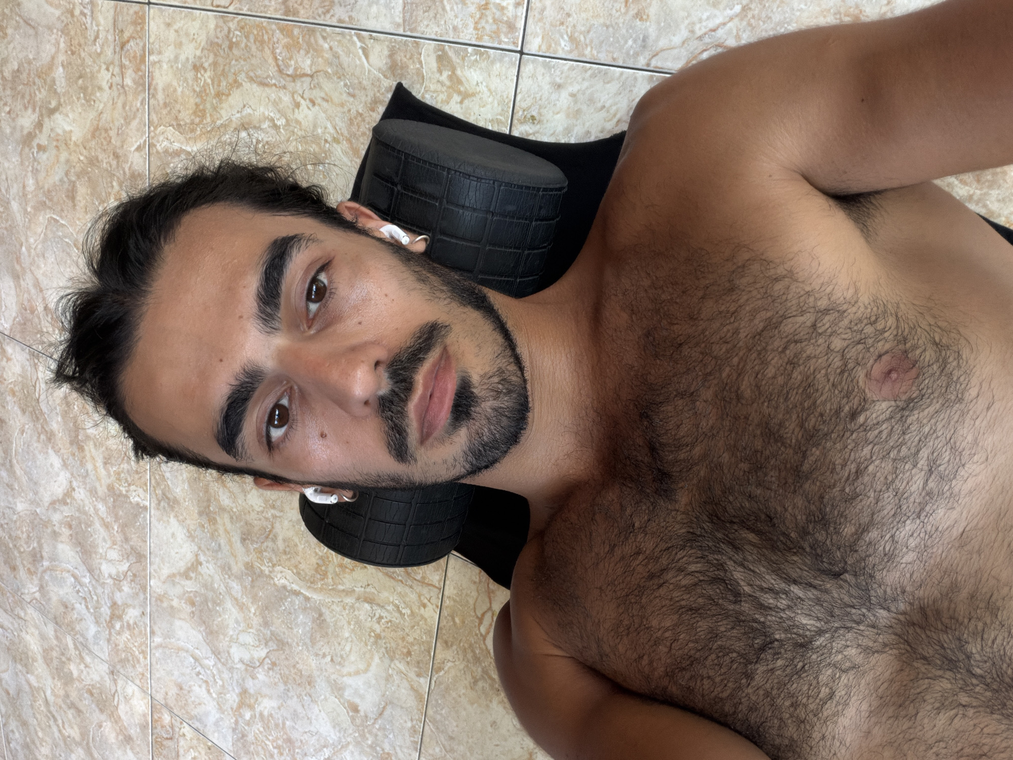 A close-up, high-angle photograph shows a shirtless man lying on a brown tile floor. He is resting his head on a black cylindrical cushion or pillow. The man has dark hair, a dark beard, and dark eyebrows. He is wearing white earbuds. The location is inferred to be North Kuta, Indonesia.