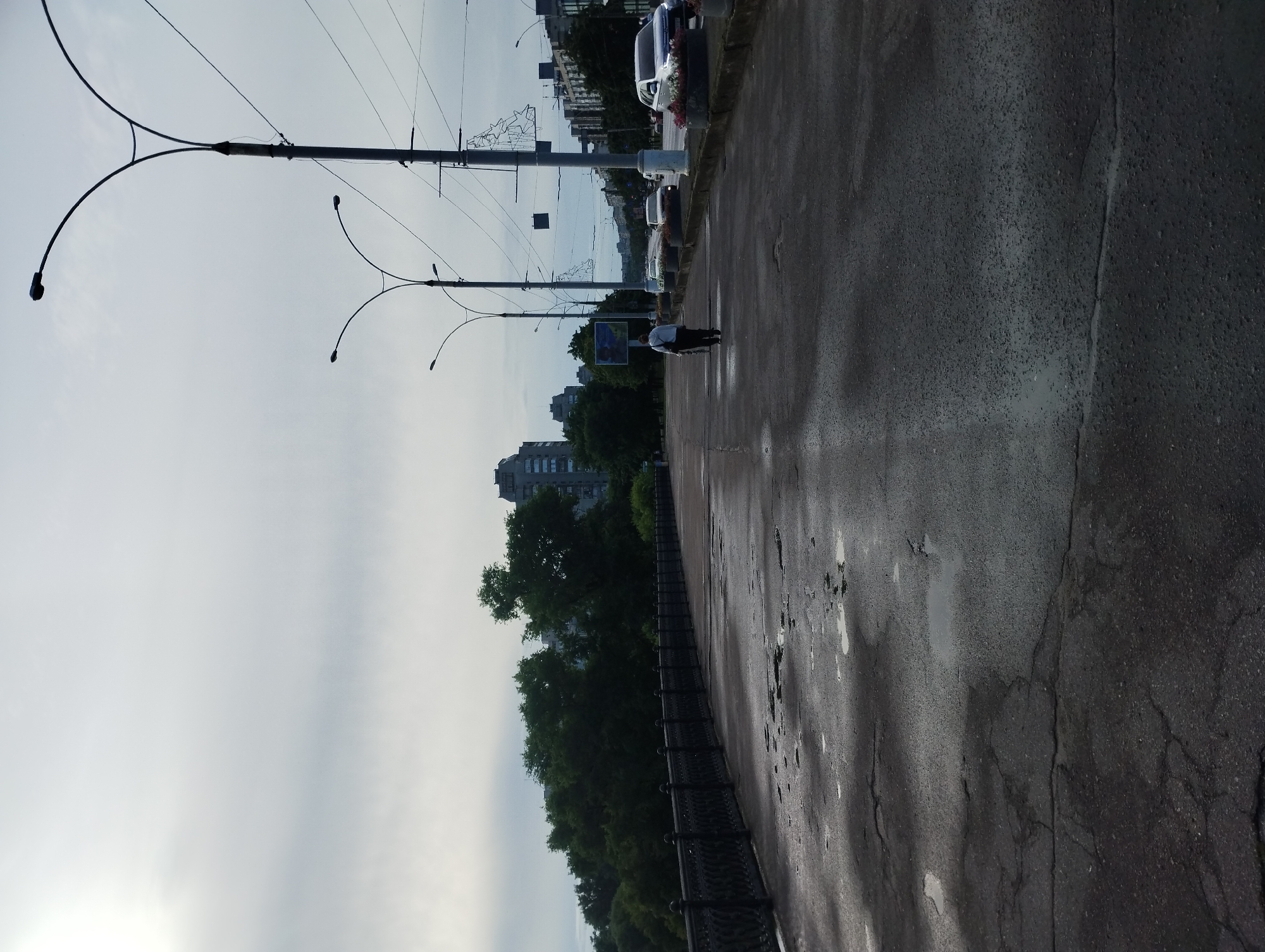The image shows a view of the embankment along the Psel River in Sumy, Ukraine. The embankment is paved with asphalt, which is wet and shows some cracks and puddles, suggesting recent rain or high humidity. A black metal railing with decorative elements runs along the left side of the embankment. Beyond the railing, there are trees and some multi-story buildings in the background.
On the right side of the embankment, there is a road with several cars driving. Streetlights with distinctive curved arms line the embankment, and overhead power lines are visible. One person is walking away from the camera on the embankment, carrying a bag. The sky is overcast and bright, with the sun partially obscured by clouds. The overall scene depicts a somewhat overcast day on a city embankment.