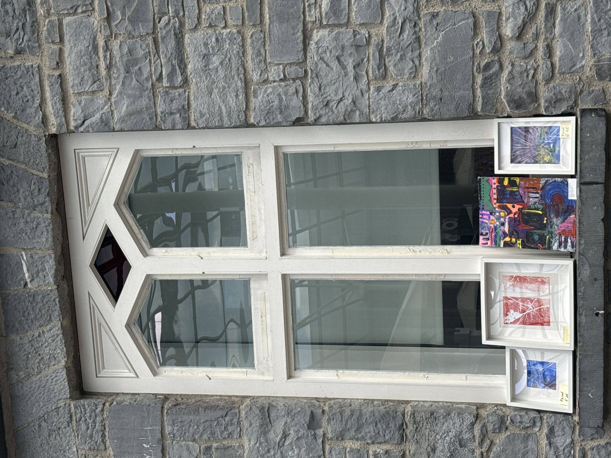 A close-up, eye-level, vertical shot shows a stone building in Kilkenny, Ireland, with a white-framed window displaying four framed artworks on the stone ledge. The window is divided into four panes, with two upper panes featuring a diamond-shaped leaded glass insert at the top. The stone facade is composed of rough-hewn grey blocks. On the ledge, from left to right, are three framed pieces of art. The first, a small square frame, contains a painting with blue and yellow tones. The second, a larger square frame, displays a red and white abstract print. Next to it is a vibrant, colorful abstract painting without a frame. To the right of this painting is a third framed artwork, a vertical rectangular frame with a landscape painting of a road leading to a city under a starry sky. The window reflects a building with a curved roof structure and lighting.