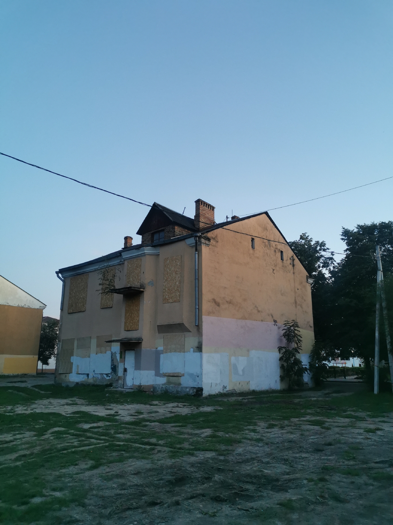 An exterior, eye-level, medium shot captures an abandoned, two-story building under a clear, pale blue sky in Brest, Belarus. The building's facade is painted in a faded peach color with peeling white and pink paint at the base. The windows on both stories are boarded up with plywood. A single tree grows from a balcony-like structure on the second floor. A small attic window is visible under the dark, sloped roof, which is topped by a brick chimney. A dark power line stretches horizontally across the sky above the building. To the left of the main building, a portion of another building, painted yellow and white, is visible. The ground in front of the buildings is covered with patchy grass and dirt. Dark green trees are located behind the building on the right. A utility pole stands to the far right of the frame, partially obscured by the trees. The scene appears to be illuminated by natural daylight, possibly during dawn or dusk due to the low angle of light.