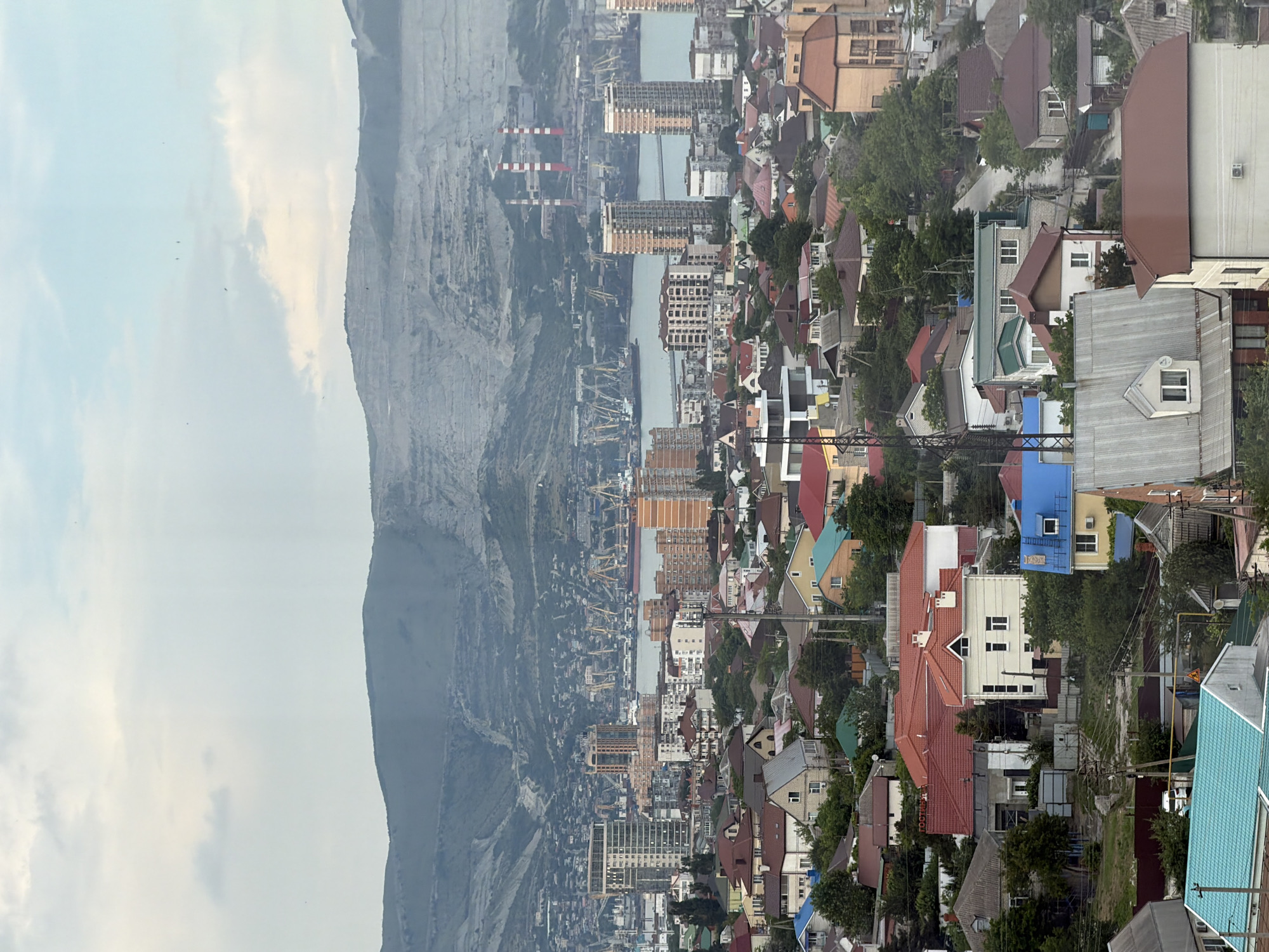 This is a high-angle, eye-level, medium shot of a residential neighborhood in Novorossiysk, Russia, with a port and mountains in the background. The sky is overcast. The foreground is dominated by a dense collection of houses with red, brown, and blue roofs. Some houses are multi-story with light-colored facades, while others are smaller, single-family dwellings. Many of the houses have visible chimneys and are surrounded by trees and greenery. In the mid-ground, there are taller apartment buildings and commercial structures. Further in the background, a large port with numerous cranes and ships is visible, situated on a body of water. Beyond the port, the landscape rises into a steep, rocky mountain range with visible quarrying activity and industrial smokestacks. The overall scene depicts a densely populated urban area transitioning into industrial and natural landscapes. No people are discernible in the image.