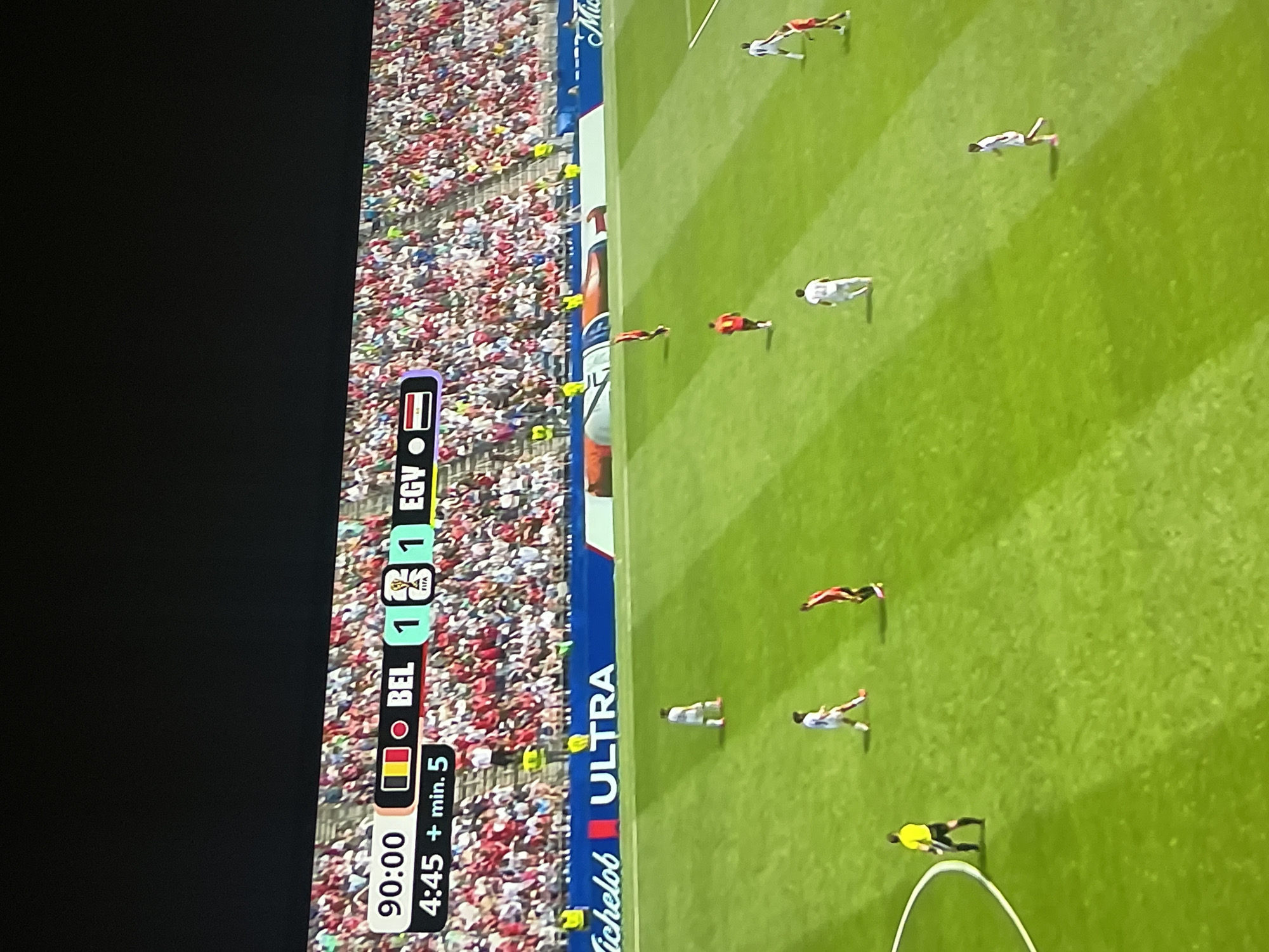 The image shows a soccer match in progress. The scoreboard indicates that the game is in the 90th minute, with added time of 4 minutes and 5 seconds. The score is tied 1-1 between Belgium (BEL) and Egypt (EGY). Several players in white and red jerseys are visible on the green field, along with a referee in a yellow jersey. The stands behind the field are filled with spectators. Commercial advertisements are visible on a blue barrier along the field, including "Michelob ULTRA." The scene is illuminated by bright daylight, suggesting it is an outdoor stadium. There is no information to suggest this location is in Ruse, Bulgaria.