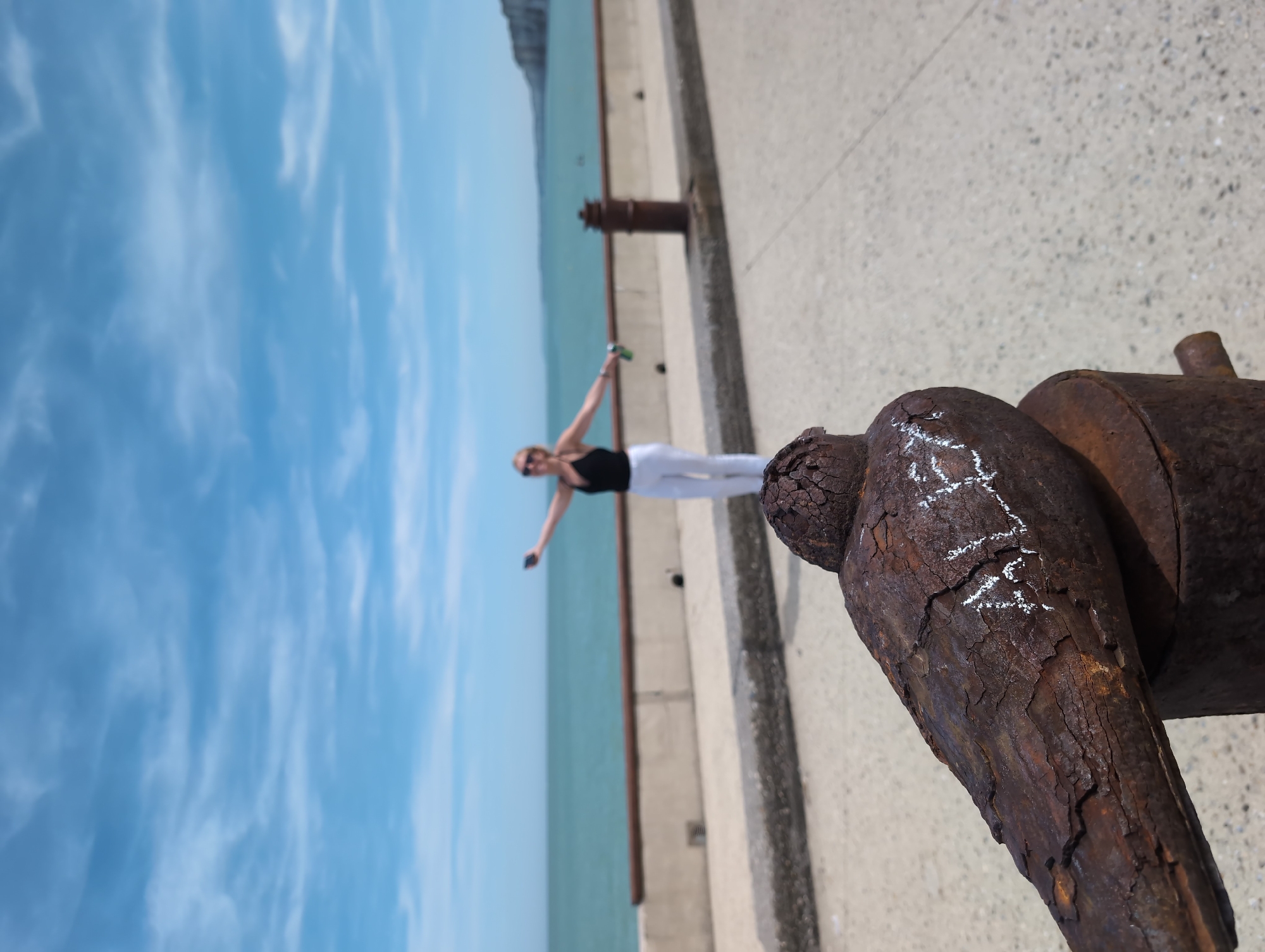 In Saint-Valery-en-Caux, France, a weathered, rusty metal object resembling a bird's head is in the foreground, with white chalk graffiti on its surface. Beyond it, a woman with blonde hair, wearing sunglasses, a black tank top, and white pants, stands with her arms outstretched, holding a green can in her left hand and a mobile phone in her right. She is positioned on a paved walkway next to a concrete wall, with the turquoise sea and a hazy coastline visible in the background under a blue sky with scattered clouds.