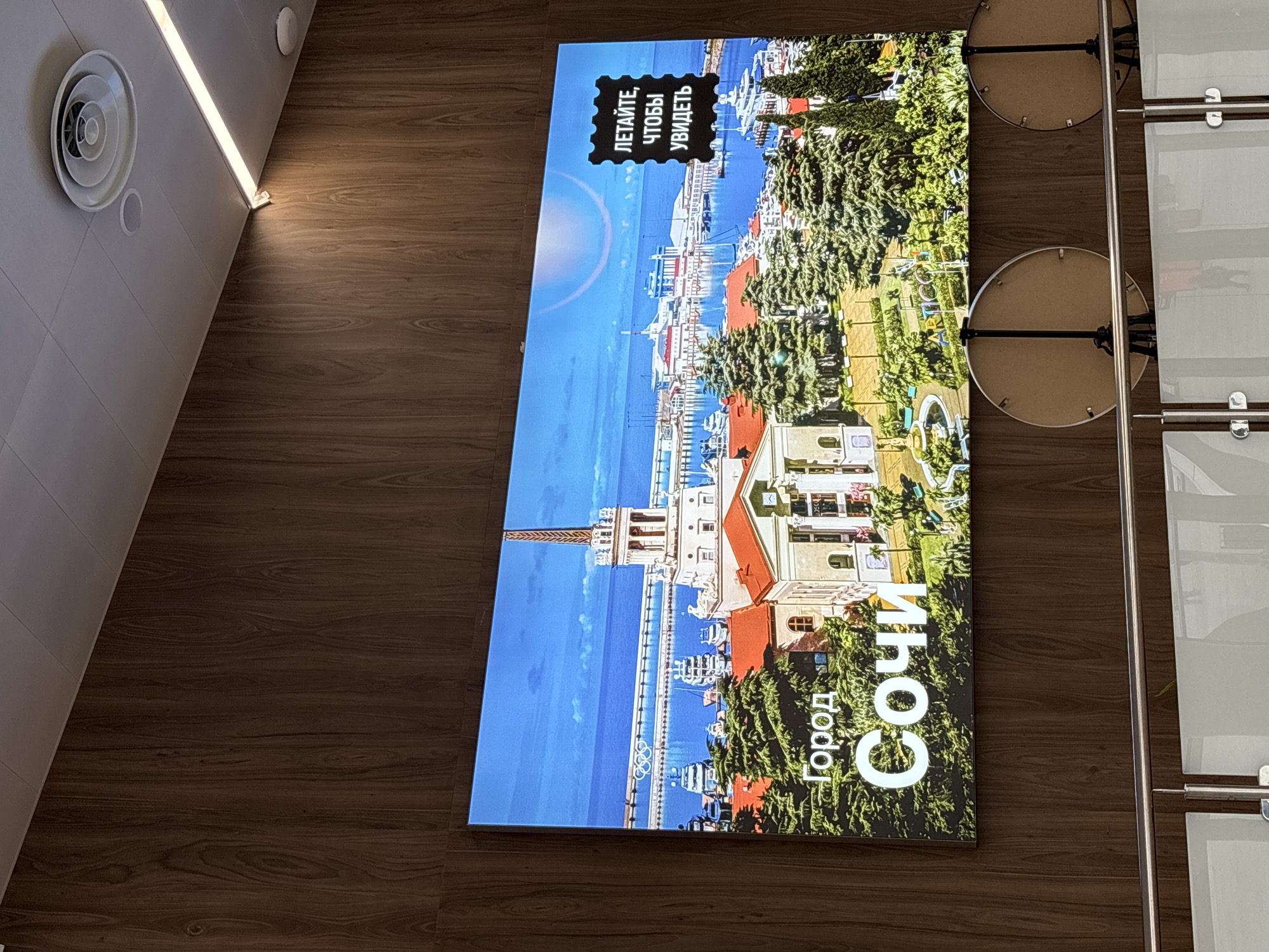 The image shows a vertical, large-format digital display mounted on a wooden wall. The display is showing a vibrant image of a coastal city with a clear blue sky and calm water. In the foreground of the display, there is lush green foliage and buildings with red roofs. A prominent architectural feature, resembling a tall tower or spire, is visible on the left side of the display. The sea is filled with yachts and boats docked along a waterfront. The Olympic rings symbol is visible in the water.
On the upper left portion of the display, there is a black stamp-like graphic containing white text in Russian. The text reads "ЛЕТАЙТЕ, ЧТОБЫ УВИДЕТЬ" which translates to "FLY TO SEE". Towards the bottom of the display, larger white text in Russian reads "Город Сочи" meaning "City of Sochi".
The overall scene depicted on the screen suggests a sunny day, likely during warmer seasons, promoting tourism or a sense of travel. The lighting on the wooden wall and the ambient light suggest the photograph was taken indoors, possibly in a lobby or public space. To the left of the display, a white circular ventilation grille is visible on a light-colored tiled surface. To the right of the display, metallic poles and glass panels suggest the presence of a modern interior design, possibly a hallway or an entrance area. The image is captured from a slightly upward-angled perspective, emphasizing the size and placement of the digital display.