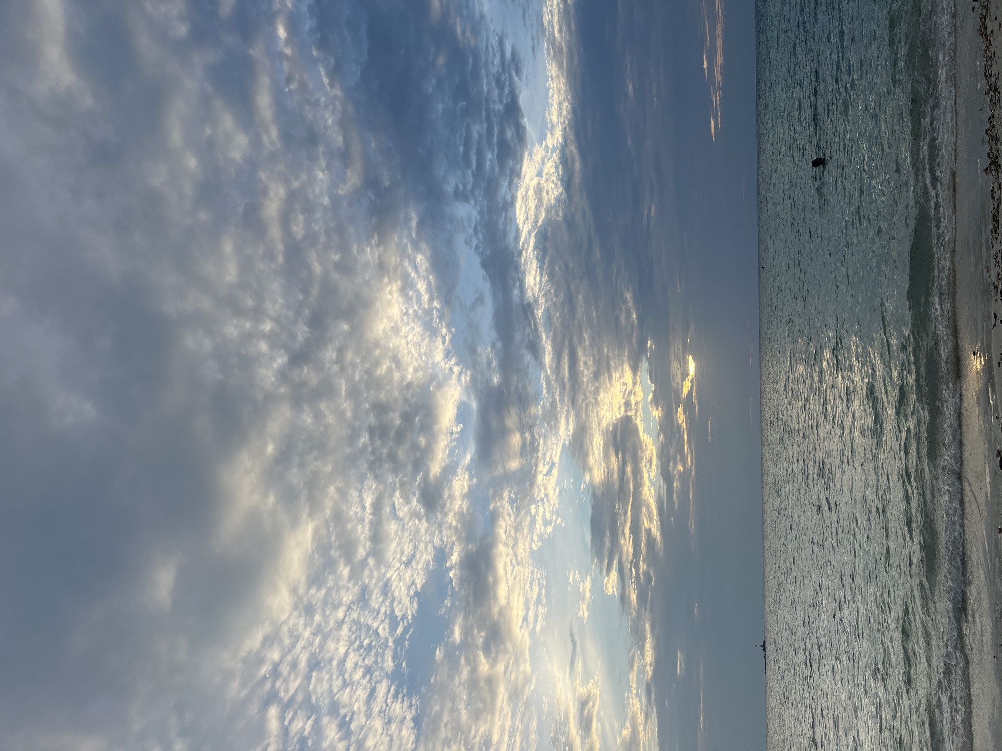 The image depicts a serene ocean scene at sunset on Ko Pha-ngan District, Thailand. The sky is filled with dramatic clouds, illuminated by the setting sun which casts a warm glow through gaps in the cloud cover. The ocean is calm, with gentle waves lapping at a sandy shore. In the distance, a small lighthouse stands on a rocky outcrop. Closer to shore, a person is swimming in the water, with only their head visible above the surface. The overall atmosphere is peaceful and tranquil.