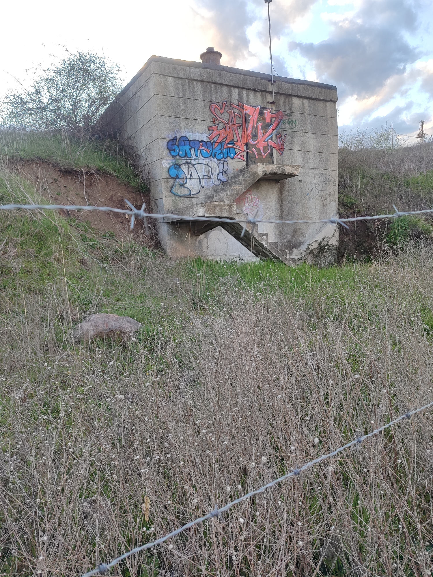 The image depicts a weathered, concrete structure with graffiti on its exterior, partially obscured by vegetation and a barbed wire fence in the foreground. The structure appears to be a small building or bunker, with an open entrance showing steps leading downwards. The surrounding environment consists of dry grass and overgrown plants on a slightly sloping terrain. The sky is overcast. The specific location is not discernible from the image, and the image does not provide enough evidence to verify it is in Burgas, Bulgaria. No people are visible.