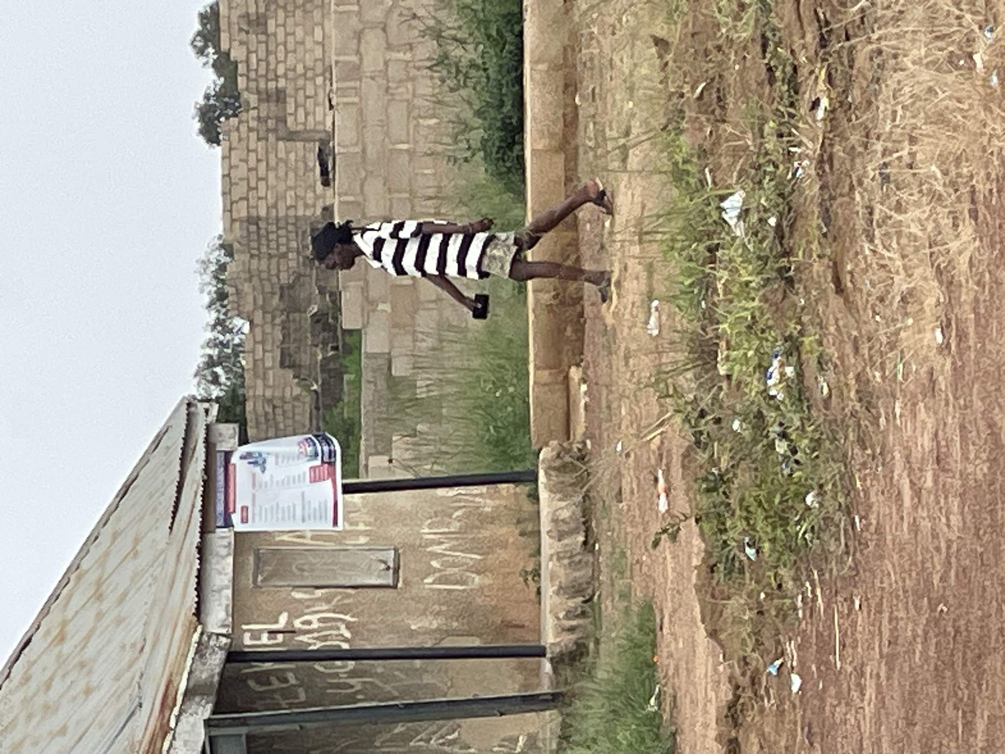 A person wearing a striped shirt and shorts walks along a dirt path in Durumi, Nigeria. The person is carrying a dark rectangular object in their right hand. To the left of the path is a small, weathered building with graffiti on its wall. A sign with text and images is attached to the front of the building. In the background, there is a partially constructed brick wall. The ground is covered with dry grass and scattered debris, suggesting an outdoor, potentially rural or underdeveloped urban environment. The sky is overcast.