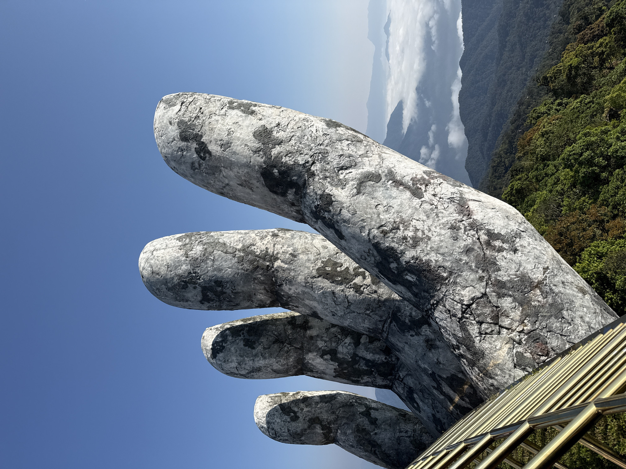 The image displays a close-up, upward-angled view of the Golden Bridge (Cầu Vàng) located in Bà Nà Hills, approximately 25 kilometers southwest of Đà Nẵng, Vietnam. The prominent subjects are four massive, weathered, gray and dark-hued stone-like fingers of a colossal hand sculpture, occupying the mid-ground and extending into the upper left of the frame. These fingers exhibit cracks and textured surfaces, simulating aged rock. In the bottom-left corner, a section of the bridge's golden railing is visible, consisting of multiple parallel horizontal bars angled upwards and to the right. The background features a clear, vibrant blue sky dominating the upper right. Below the sky and beyond the hand, a panoramic mountainous landscape unfolds, characterized by dense dark green forests. A distinct cloud inversion is present, with a thick layer of white, fluffy clouds resembling a sea nestled between mountain peaks in the middle distance. The scene is illuminated by bright natural light, indicating a clear day. No human figures are present.