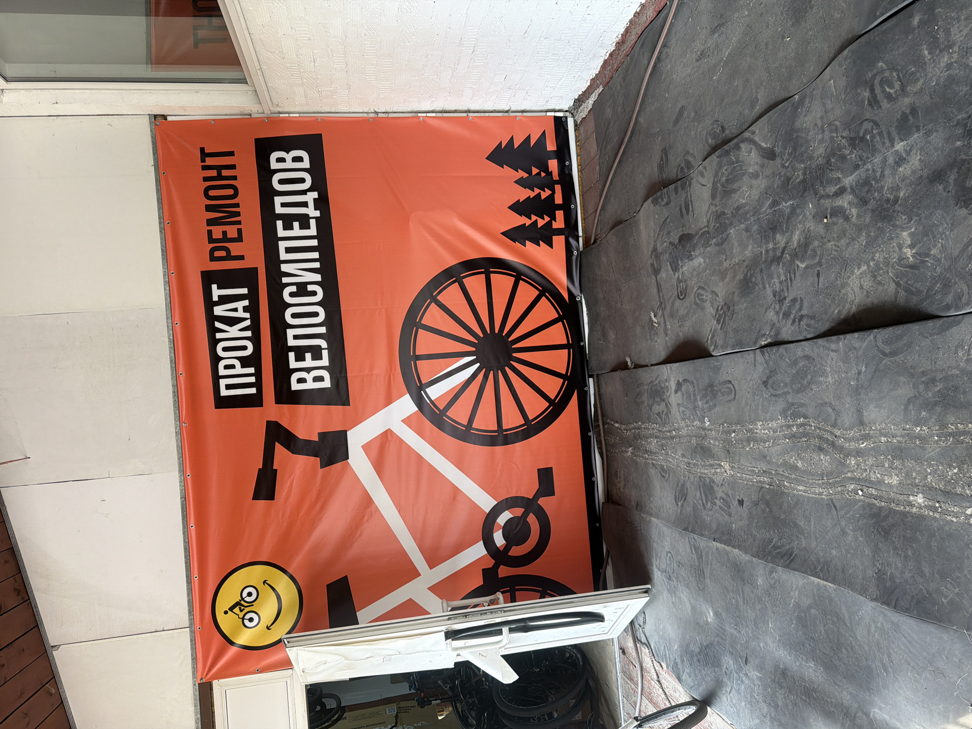 The image shows a large orange banner with black text and graphics, likely advertising a bicycle rental and repair shop. The banner features a cartoon smiley face with bicycle wheels for eyes, a stylized bicycle frame, and silhouettes of pine trees. The text on the banner reads "ПРОКАТ РЕМОНТ ВЕЛОСИПЕДОВ" (Rental Repair Bicycles). The banner is hanging outside a building, possibly a garage or workshop, with a white door open to the left. The ground in front of the banner is covered with dark, textured material, possibly rubber mats or asphalt, showing tire tracks and dust. In the background, through the open doorway, several bicycles are visible. The overall scene suggests an outdoor commercial space in an urban or industrial area. The mention of Novosibirsk, Russia, places this scene within that specific geographical context.