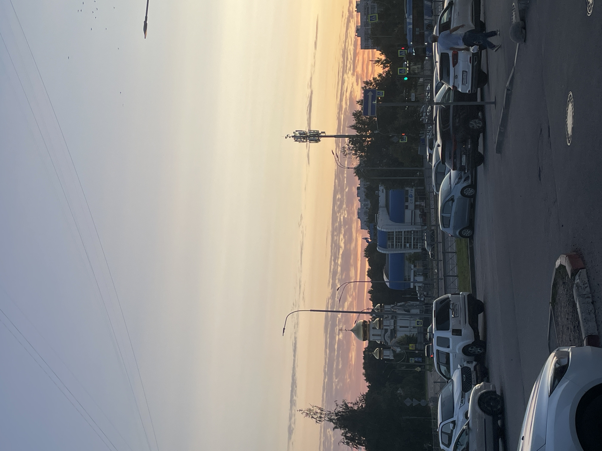 The image depicts a street scene at dusk in Saint Petersburg, Russia. A white SUV is parked in the foreground, facing away from the camera. Behind it, a black SUV is also parked, and further back, a white sedan and a silver sedan are visible. In the middle ground, a white church with golden domes stands prominently, surrounded by trees. To the right of the church, a modern building with blue accents is situated. A cell tower is visible to the right of the church and building. A person wearing a blue shirt and jeans is walking away from the camera in the middle right of the frame. The sky is a gradient of pale blue and orange, with clouds visible. Power lines crisscross the sky. The scene suggests a typical urban environment with traffic and religious architecture.