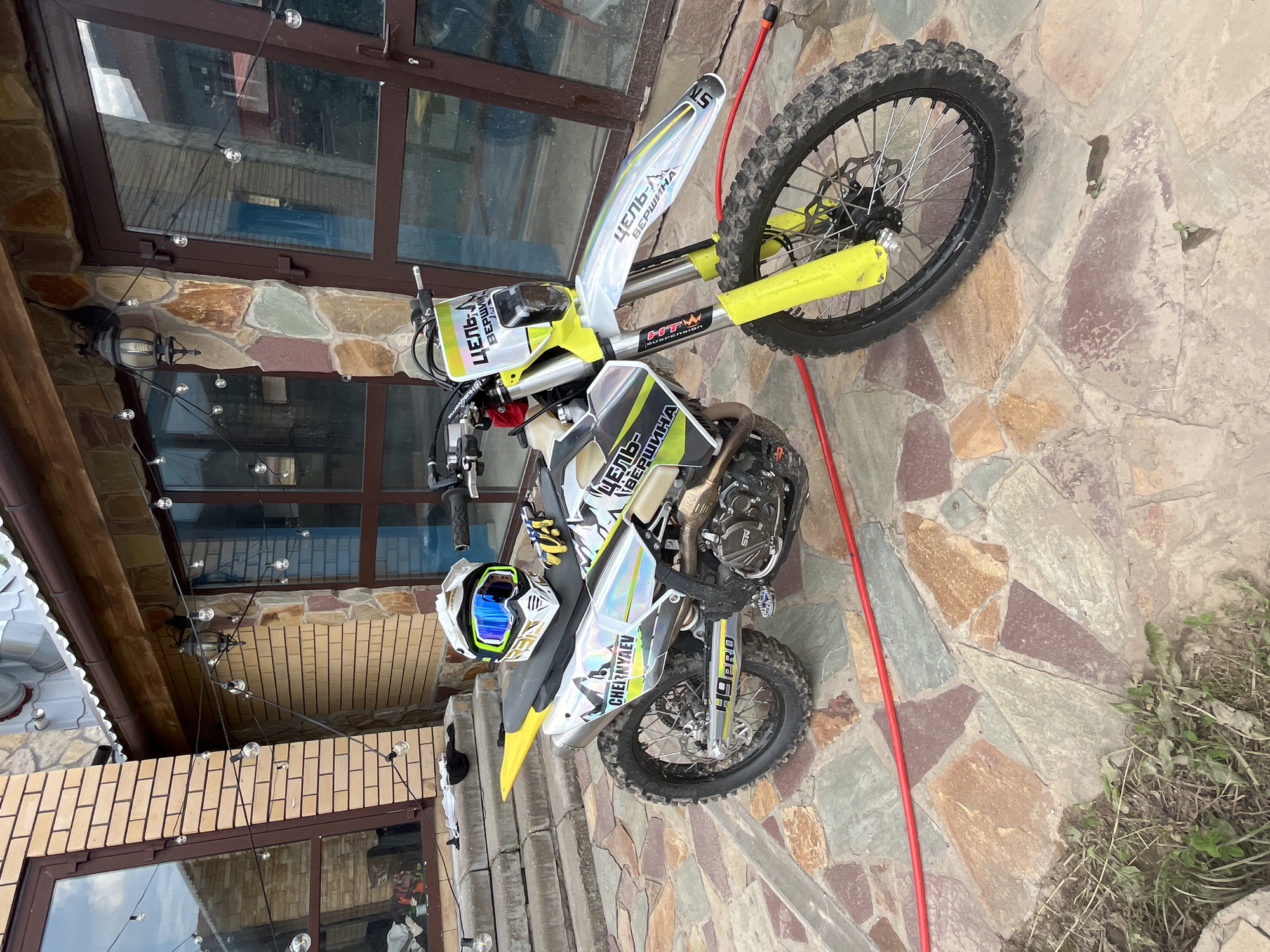 A white and yellow dirt bike with blue and green goggles on the seat is parked on a flagstone patio in front of a house in Orekhovka, Russia. The house has a brick and stone facade and large windows. A string of globe lights hangs across the front of the house. A red hose lies on the ground near the motorcycle.