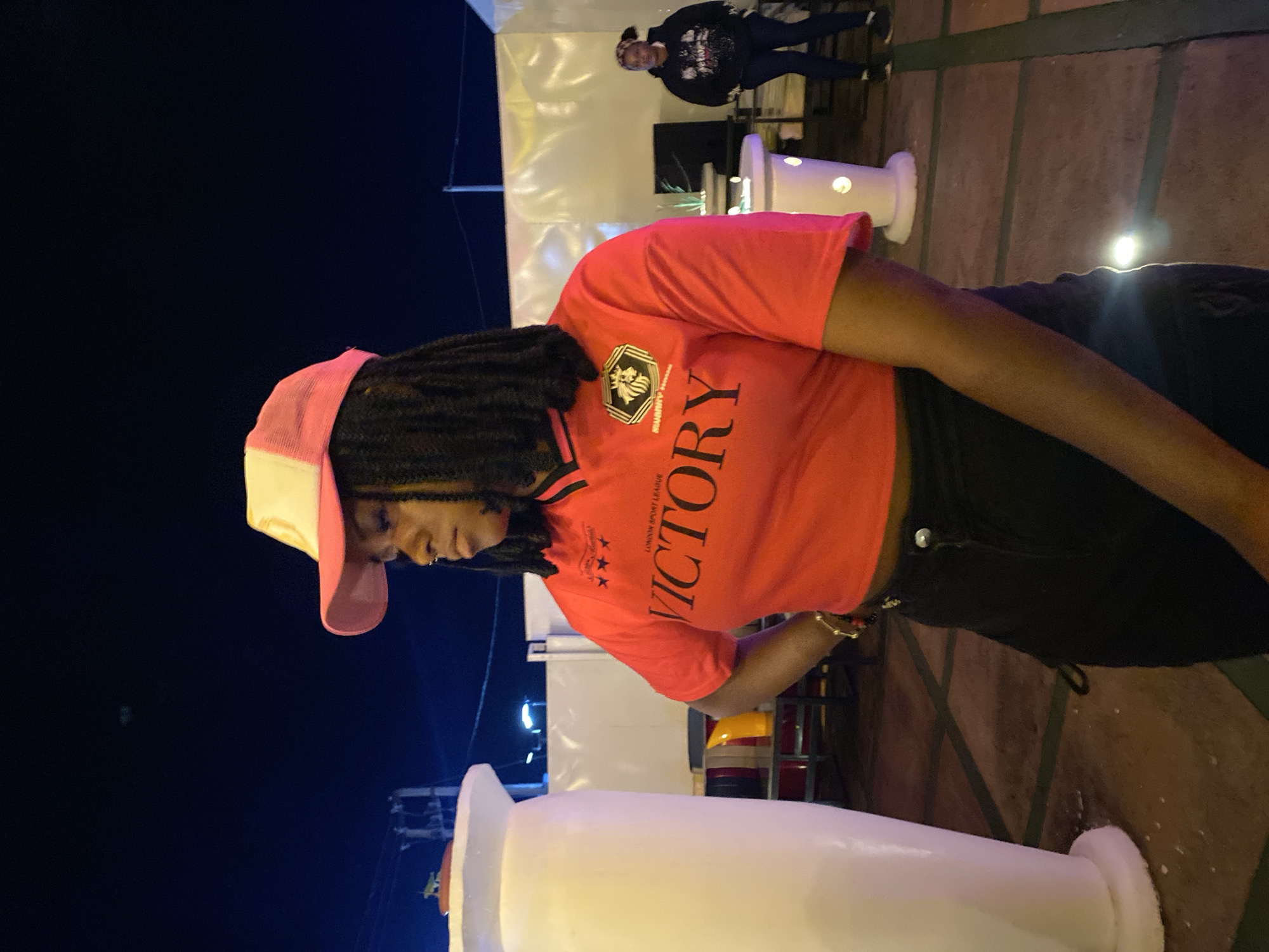 A young Black woman wearing a red shirt with "VICTORY" printed on it and a pink and white baseball cap stands with her left hand on her hip. Her dark hair is styled in dreadlocks. She is wearing black pants. The background shows an outdoor setting at night with white tent-like structures and illuminated white pillars. Another person is visible in the background. The ground appears to be paved with dark tiles and some lighter colored lines. The overall lighting suggests an artificial light source.