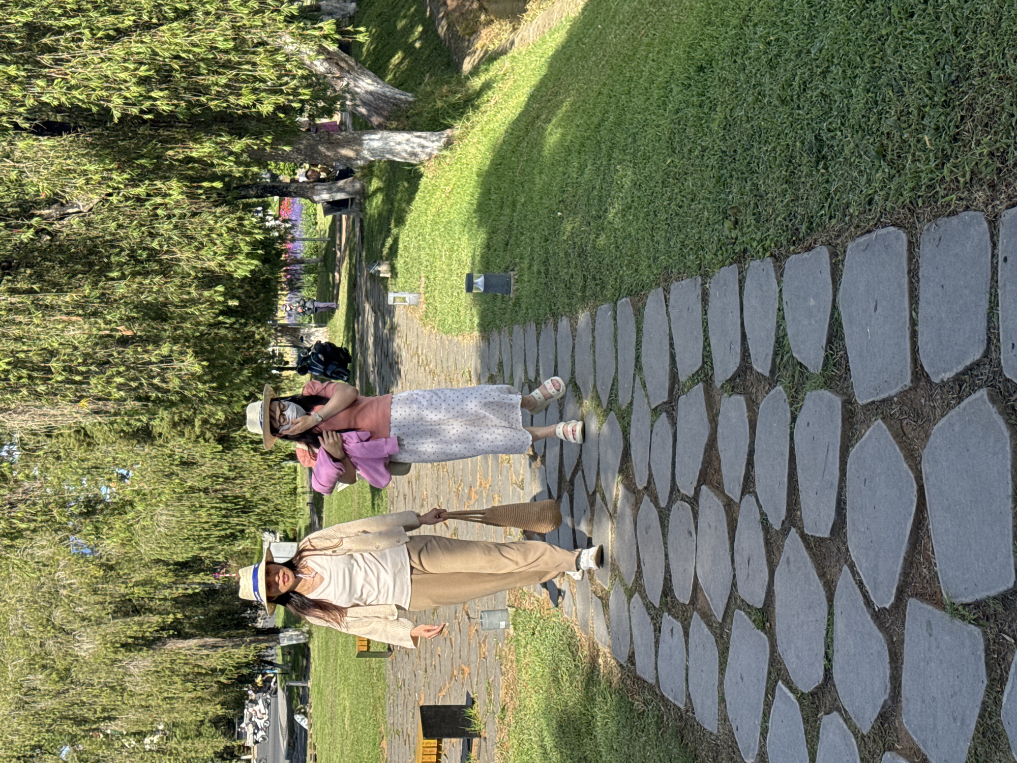 In Da Lat, Vietnam, two women are walking on a stone pathway in a park. The woman in the foreground wears a straw hat, a beige cardigan over a cream-colored top, and tan pants, carrying a woven tote bag. The woman behind her, also wearing a straw hat, has a mask over her face, a pink top, and a white patterned skirt, holding a pink jacket. The pathway is bordered by lush green grass and shaded by large trees with drooping branches. In the background, there are motorbikes, other people, and a body of water.