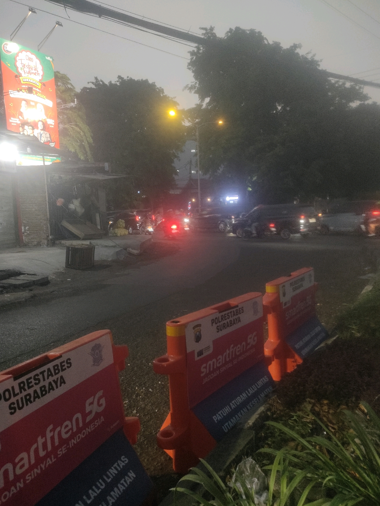 The image depicts a street scene in Surabaya, Indonesia, at dusk or dawn. In the foreground, orange traffic barriers with white and red signage are positioned along the roadside. One barrier prominently displays "POLRESTABES SURABAYA" and "smartfren 5G," along with the slogan "JAGOAN SINYAL SE-INDONESIA" and "PATUHI ATURAN LALU LINTAS UTAMAKAN KESELAMATEN." Another barrier shows "POLRESTABES SURABAYA" and "smartfren 5G."
Further down the street, traffic is visible, including cars and motorcycles with illuminated taillights. A large billboard advertising "Cleo Festival Kuliner SURABAYA" with images of people is illuminated on the left side of the frame. There are buildings and trees lining the street. The sky is overcast, and streetlights are visible. A wooden object, possibly a crate or bin, is on the sidewalk. The overall atmosphere suggests an urban environment with ongoing traffic and commercial activity.