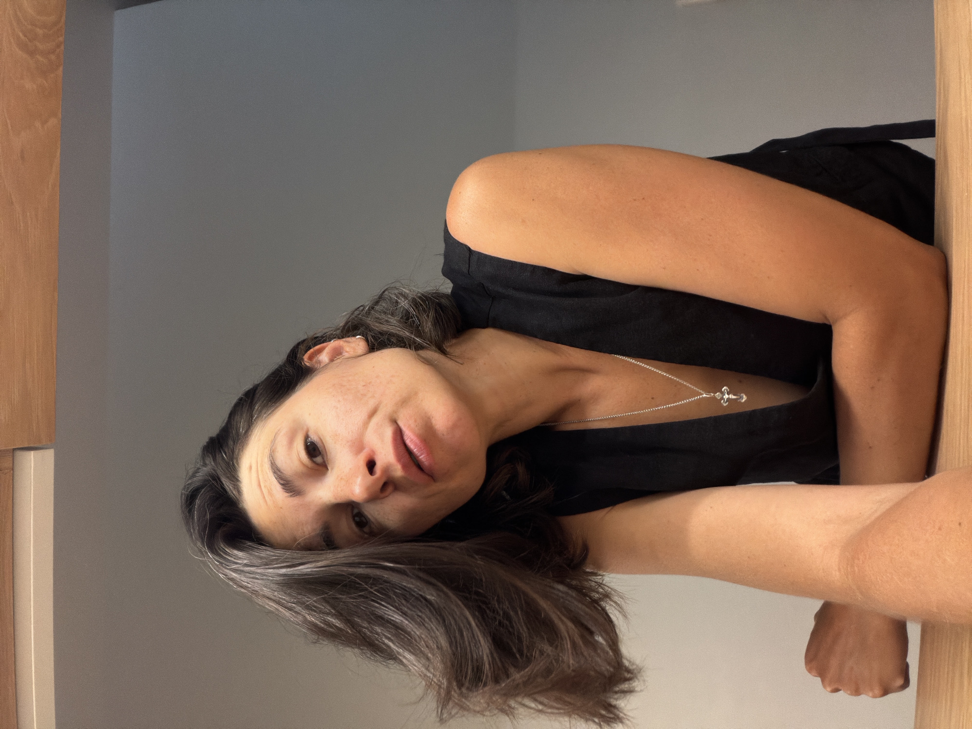 The image features a woman looking directly at the camera with a tilted head, suggesting a close-up selfie or portrait. She has dark, shoulder-length hair with visible grey strands, brown eyes, and freckles on her face. Her lips are slightly parted, conveying a relaxed or contemplative expression. She is wearing a dark, possibly black, sleeveless top or dress. A thin silver chain necklace with a small, intricate silver cross pendant hangs around her neck. Her arms are visible, suggesting she is leaning or resting them.
The setting is indoors, with a plain grey wall forming the primary background. On the left and right edges of the frame, light-colored wooden surfaces are visible, possibly parts of a door frame, wall paneling, or furniture. The lighting appears soft and even, highlighting her features. The overall impression is casual and personal. No text is visible in the image. The location context provided is Budva, Montenegro.
