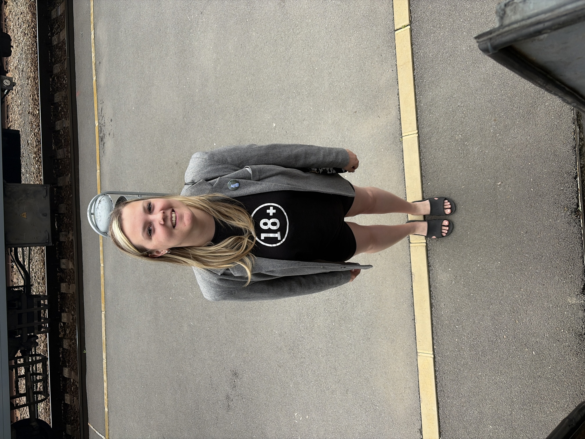 A blonde woman wearing a gray blazer, a black t-shirt with a white "18+" logo, and black sandals is standing on a paved platform next to a train track. She is facing the camera with a slight smile. The platform edge has a yellow safety line. The train carriage is partially visible on the left, and the edge of another object is visible on the right. The photo was taken outdoors during daylight. It is unlikely that the location of Rostov-on-Don can be confirmed from the image, as the scene is generic and lacks any distinctive landmarks.