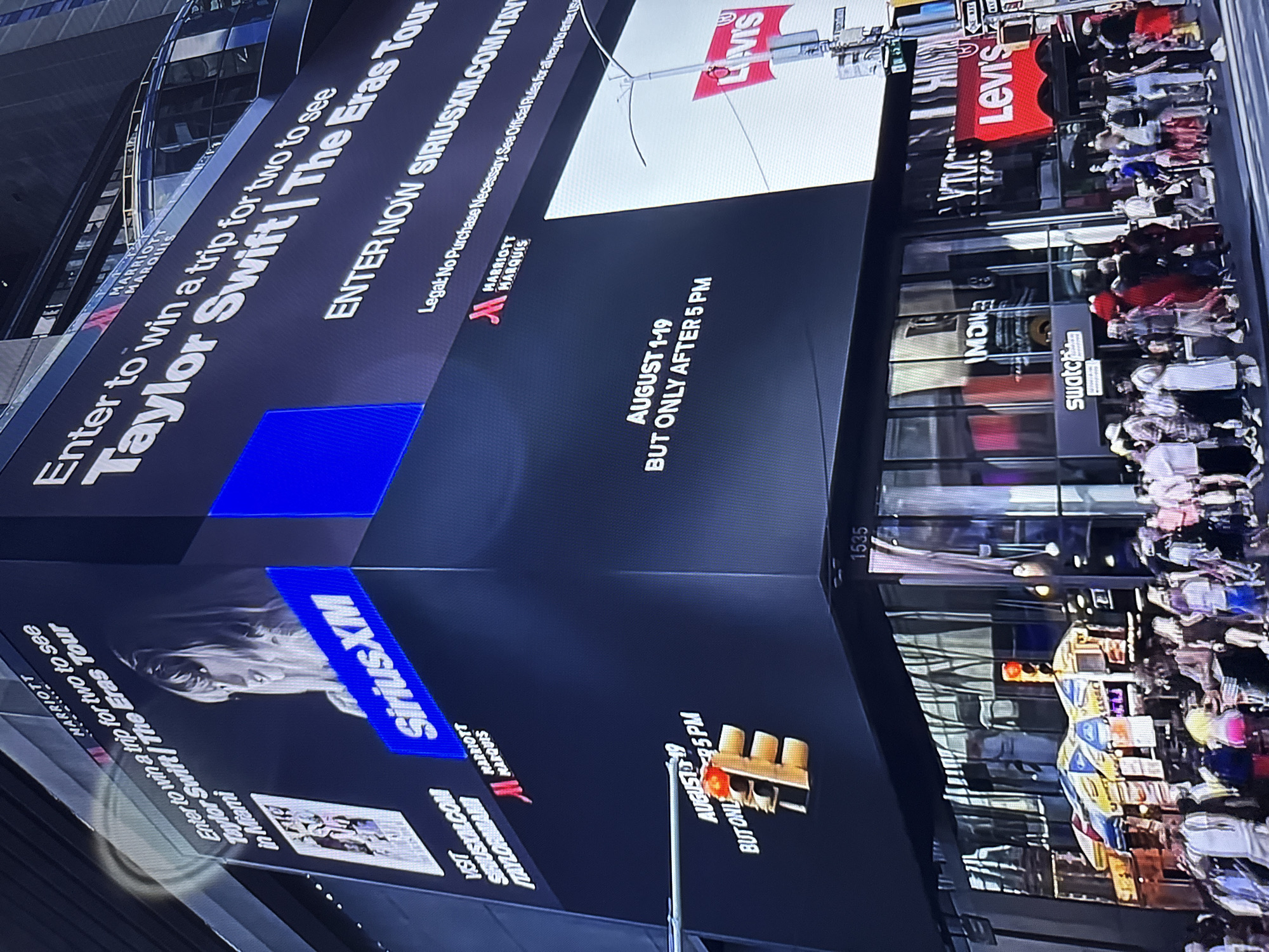 The image is a photograph taken from street level, looking up at a large, multi-story building displaying digital advertisements. The scene appears to be Times Square, New York City, given the presence of multiple large digital billboards, bright lights, and a crowded pedestrian area. The main focus is a large Marriott Marquis advertisement for a Taylor Swift "The Eras Tour" giveaway.
The billboard prominently features the text "Enter to win a trip for two to see Taylor Swift | The Eras Tour in Miami". Below this, it says "ENTER NOW SIRIUSXM.COM/TAY". Another section of the billboard, facing a different angle, advertises "AUGUST 1-19 BUT ONLY AFTER 5 PM". A smaller screen on the left side shows a poster-like graphic with "VISIT SIRIUSXM.COM/TAYLORMIAMI". The Marriott Marquis logo is visible on the billboards.
Below the digital billboards, the ground level of the building has storefronts. Visible signage includes "Swatch" and "Levi's". A red Levi's logo is clearly displayed on one of the storefronts.
The street in front of the building is bustling with pedestrians, many of whom are blurred due to motion. People are dressed in casual summer attire. Several street vendors with umbrellas selling merchandise are visible. Traffic lights are also visible. The overall environment is vibrant and commercial, characteristic of a major urban entertainment district.
There is no indication that this image is located in Burgas, Bulgaria.