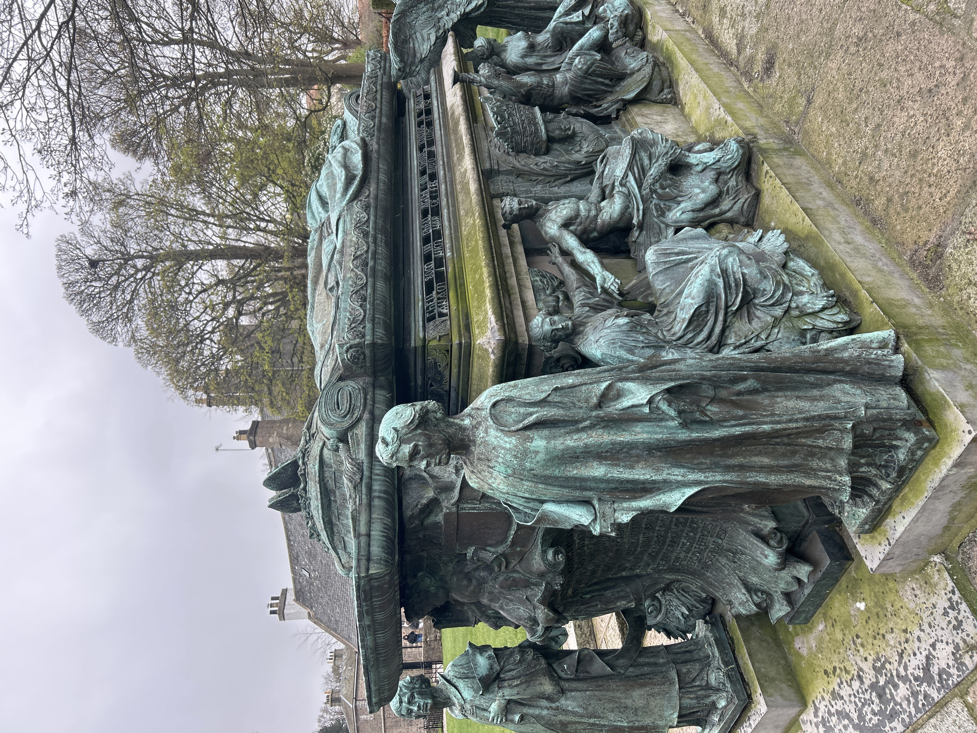 The image shows a close-up, tilted view of a bronze monument, located in Aberdeen City, United Kingdom. The monument features multiple figures, likely representing a historical or allegorical scene. The figures are draped in clothing and possess intricate detailing. The bronze has a green patina, suggesting age and weathering. The monument rests on a stone base, which is covered in moss and lichen. In the background, bare trees and a building with a grey roof and chimneys are visible. The sky appears overcast.