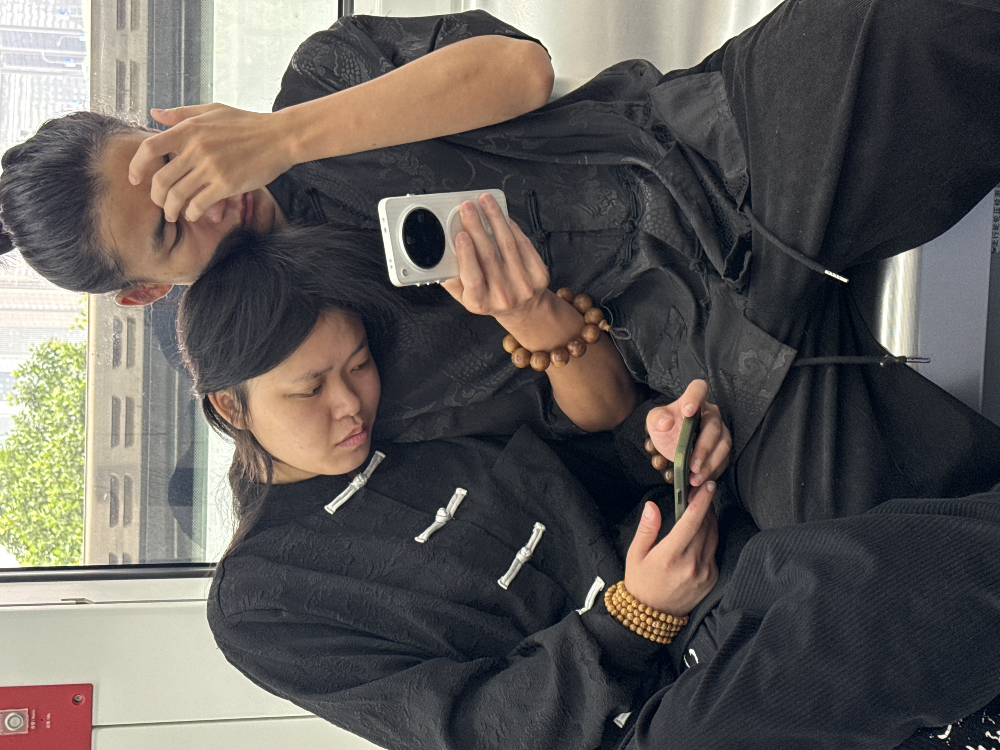 Two people, dressed in matching black traditional Chinese clothing, are seated on what appears to be a train. The individual behind is shielding their eyes with one hand and holding a silver camera in the other. The person in front is looking at a smartphone, also held in their hand. Both are wearing wooden beaded bracelets. Through the train window, a building and trees can be seen, suggesting they are above ground. Based on the "报警/报警" text near the emergency alarm button, it appears to be in Yuzhong District, China.