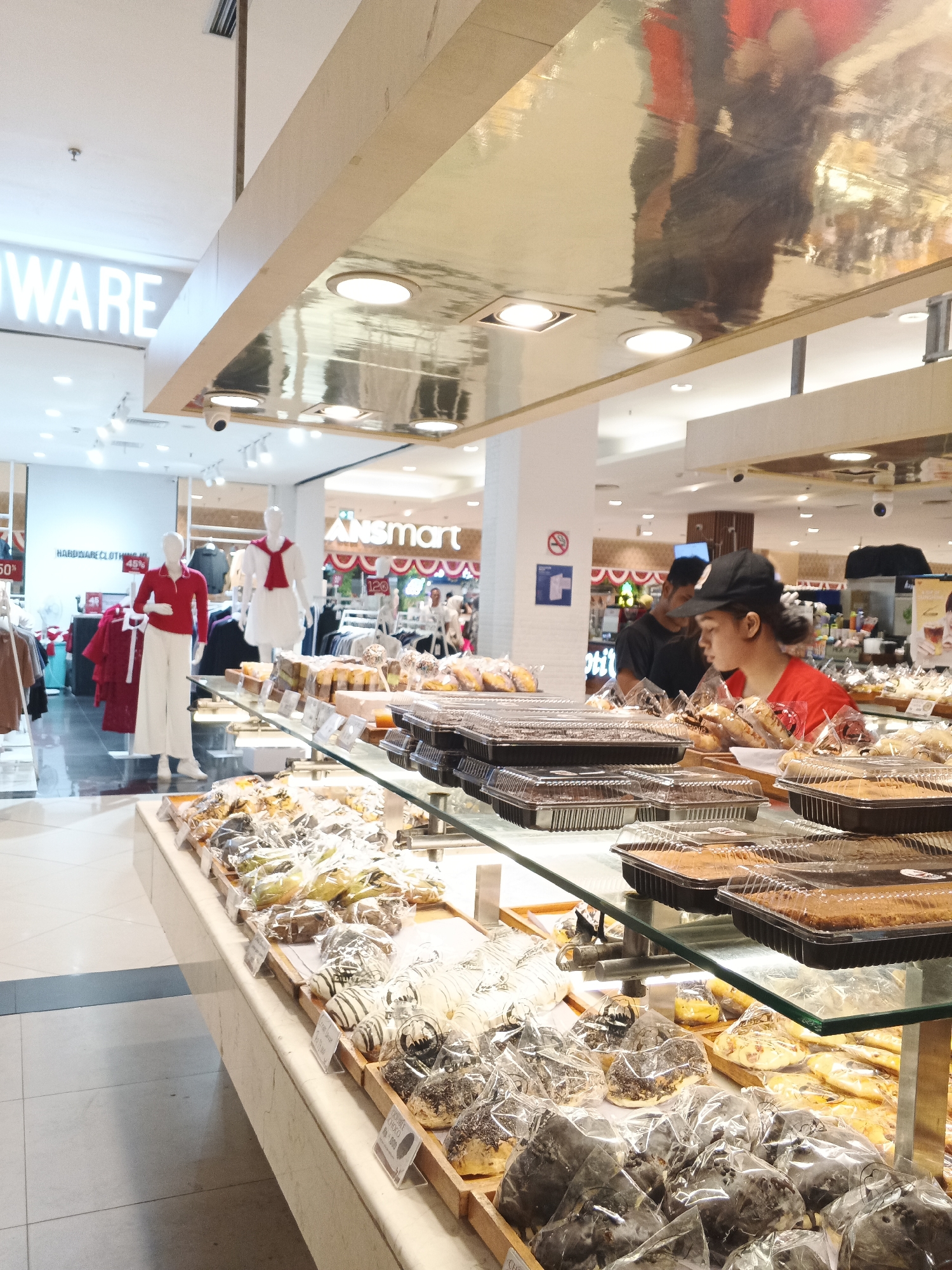 The image shows the interior of a bakery, likely located in a supermarket or department store in the City of Medan, Indonesia. In the foreground, a glass display case showcases an assortment of baked goods, including donuts, pastries, and cakes, many of which are individually packaged in clear plastic. Behind the display, a young woman wearing a red shirt and a black baseball cap is actively serving customers or arranging products, with another person, possibly a customer, visible behind her.
Further in the background, mannequins dressed in red and white clothing are displayed, suggesting the presence of a clothing section within the same establishment. Signs with text, including "HARDWARE CLOTHING" and what appears to be "Unismart", are visible on the walls, indicating a retail environment. The overall lighting is bright, with overhead lights reflecting off a metallic ceiling. The floor is tiled, with a gray and white pattern. The scene captures a typical retail setting where food and other goods are sold.