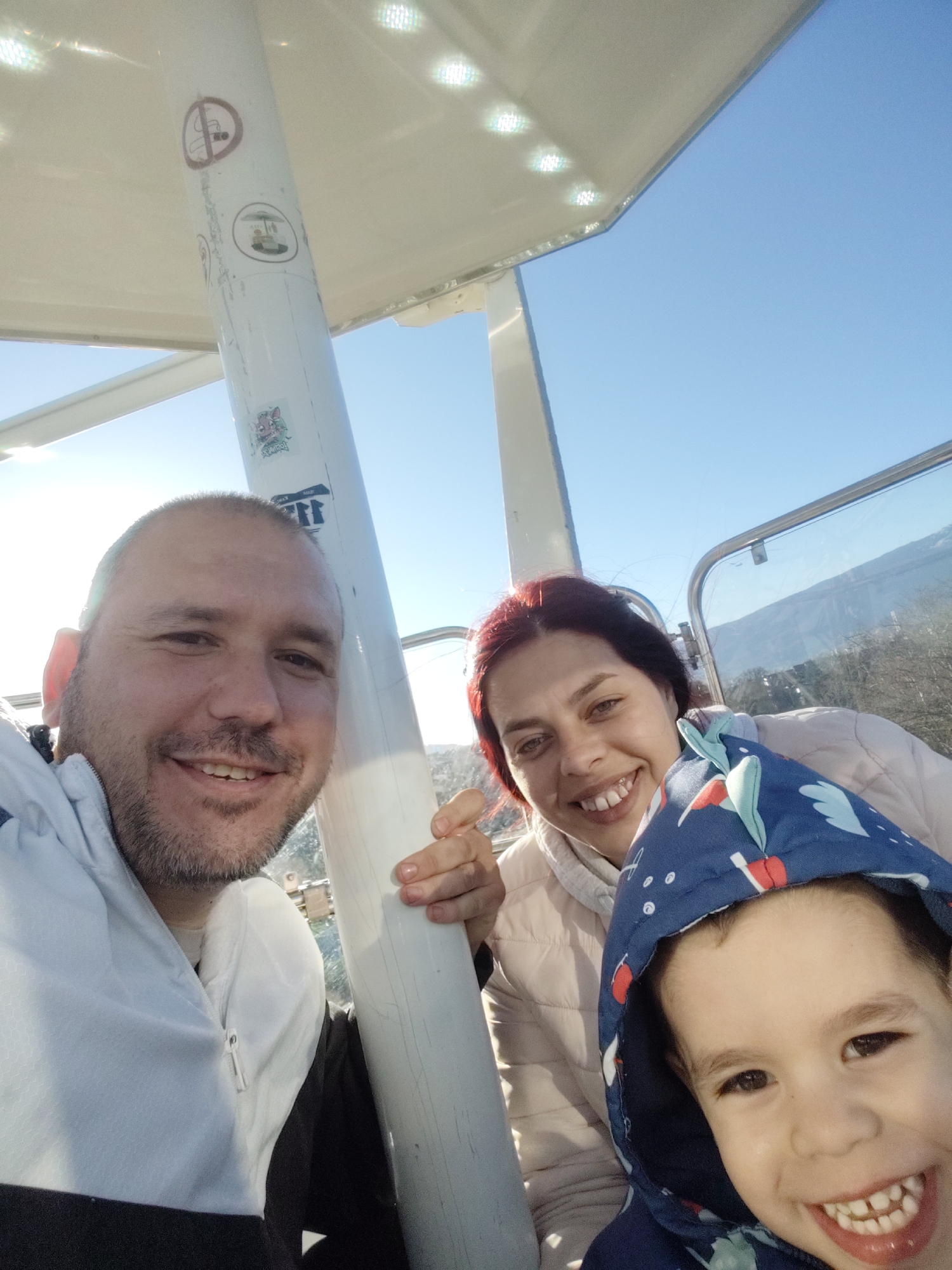 A selfie photograph captures three people in a cable car setting with a mountainous landscape visible through the car window. A man with short hair and a beard is on the left, smiling. A woman with reddish hair is positioned behind him, also smiling. A child wearing a hooded jacket is in the front, displaying a big smile. The inside of the cable car has a white structural support with safety and informational stickers attached. The background shows a clear blue sky. This scene is likely in Sofia, Bulgaria, given the mountainous landscape.