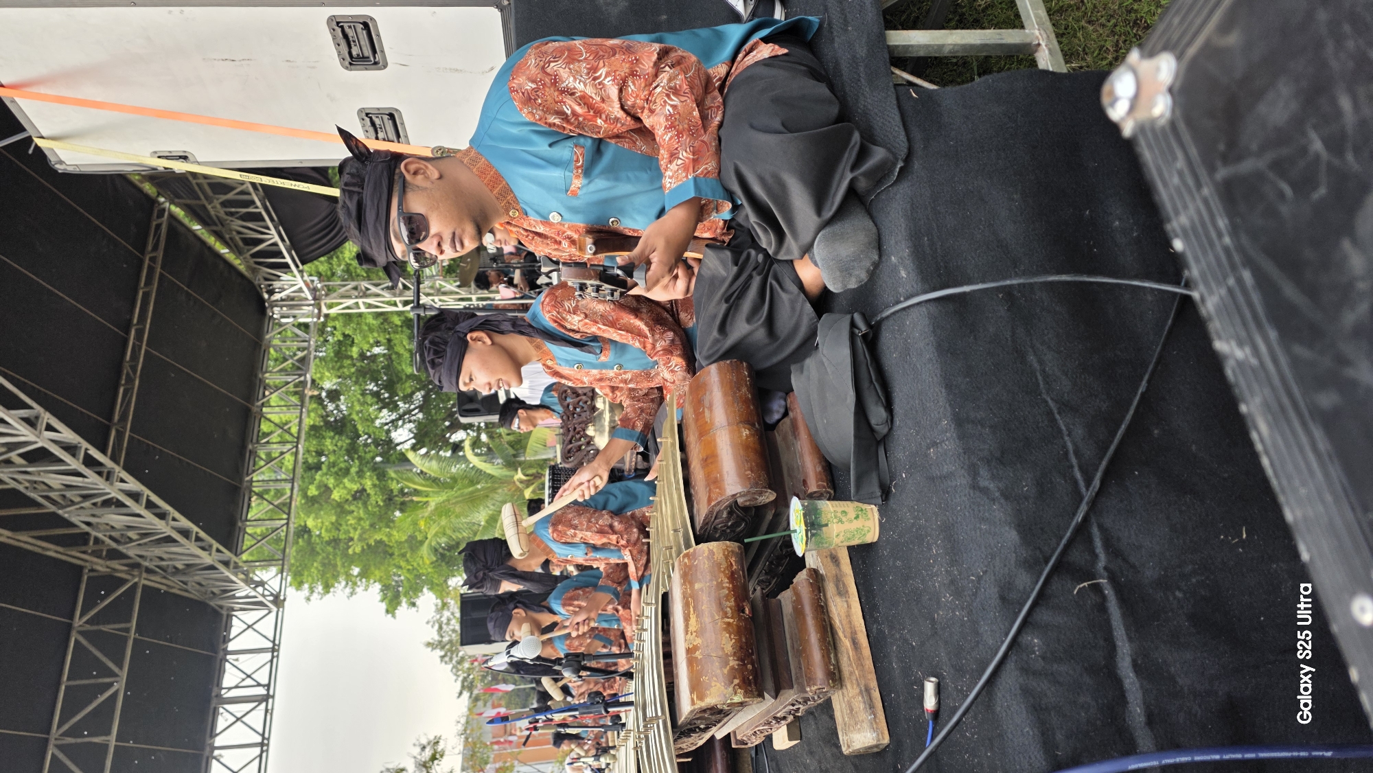A group of musicians, likely performing a traditional Indonesian Gamelan piece, are seated on a black stage beneath a metal truss structure. The musicians are dressed in matching teal and orange patterned shirts and black pants, with black headwraps. Some are actively playing instruments, including what appear to be xylophone-like percussion instruments with wooden frames and metal keys, and one musician is playing a stringed instrument resembling a ukulele or guitar. In the foreground, a partially visible cup with a straw sits on a wooden plank. The background shows green trees and a cloudy sky. The lower right corner of the image displays the text "Galaxy S25 Ultra". The scene is outdoors, possibly at an event or festival in Kecamatan Pati, Indonesia, given the presence of the phone's watermark which often indicates the location of capture.