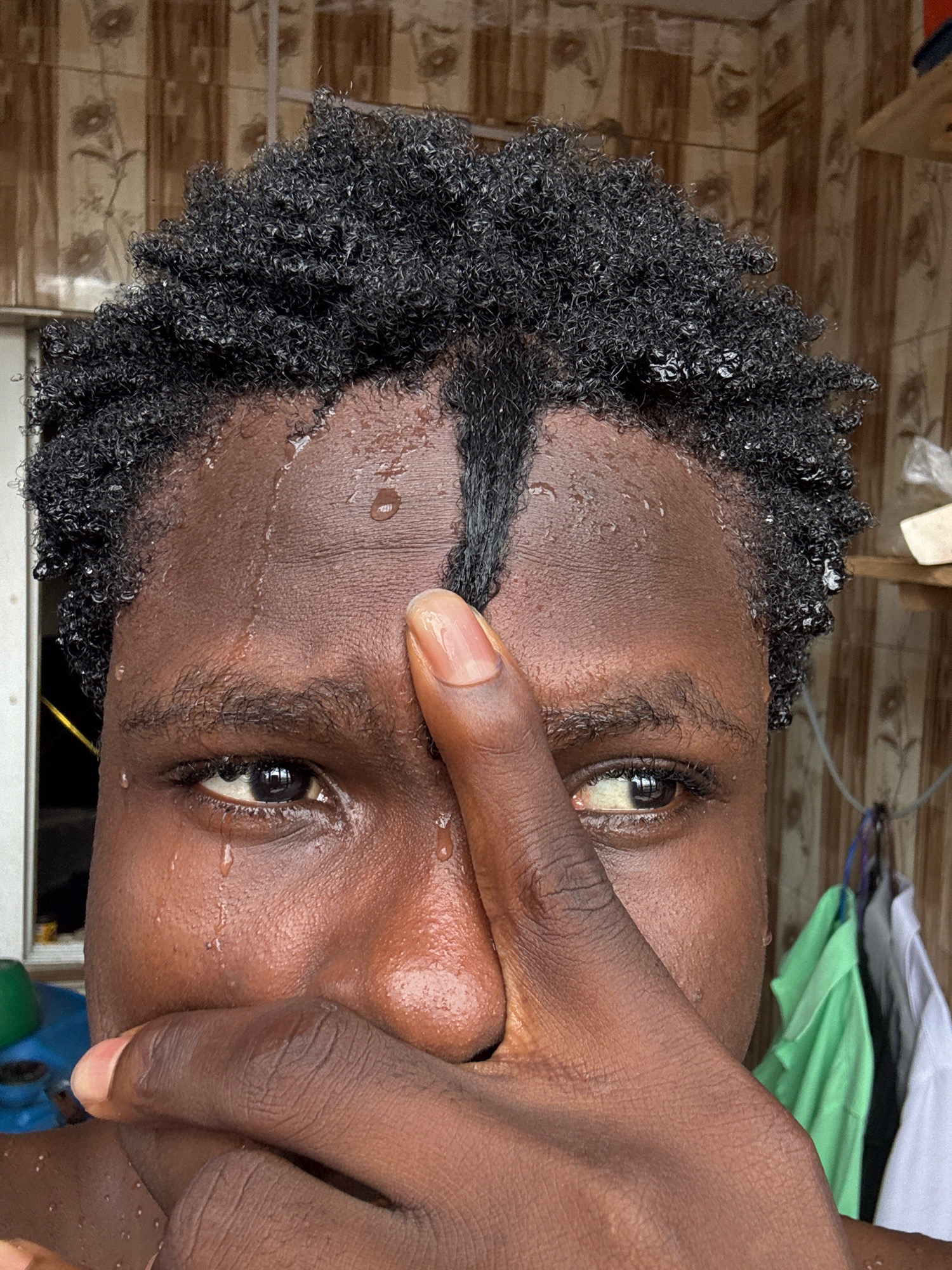 The image is a close-up, frontal view of a young person's face, with their hand partially obscuring their mouth and nose. The person has dark, curly hair and dark skin, with visible water droplets on their forehead, face, and hair, suggesting they are sweating or have recently been wet. Their eyes are open, and there are teardrop-like water droplets on their cheeks. The background appears to be a tiled wall, and on the right side, several articles of clothing are hanging. The overall impression is one of distress or intense physical exertion. The image is located in Ihiagwa, Nigeria.