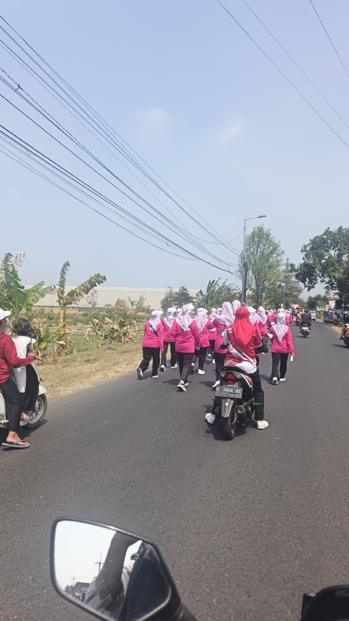 A group of women in pink tops and white or light purple headscarves are walking down a paved road. One woman in the foreground is riding a motorcycle. To the left of the road are banana trees and a building in the background. The sky is clear and blue. This scene appears to be in Mlaten, Indonesia.