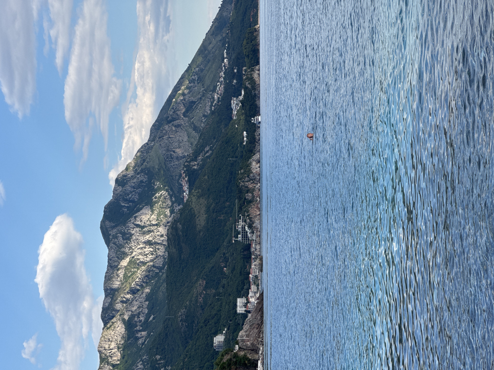 The image shows a view in Rafailovići, Montenegro. The Adriatic Sea occupies the right half of the frame, its surface rippled by wind. A small buoy or marker is visible in the water. On the left, a steep, rocky mountain covered in vegetation rises. Buildings and structures are built on the slopes, indicating residential or commercial development. The sky is visible in the top left, featuring patches of blue and white clouds.