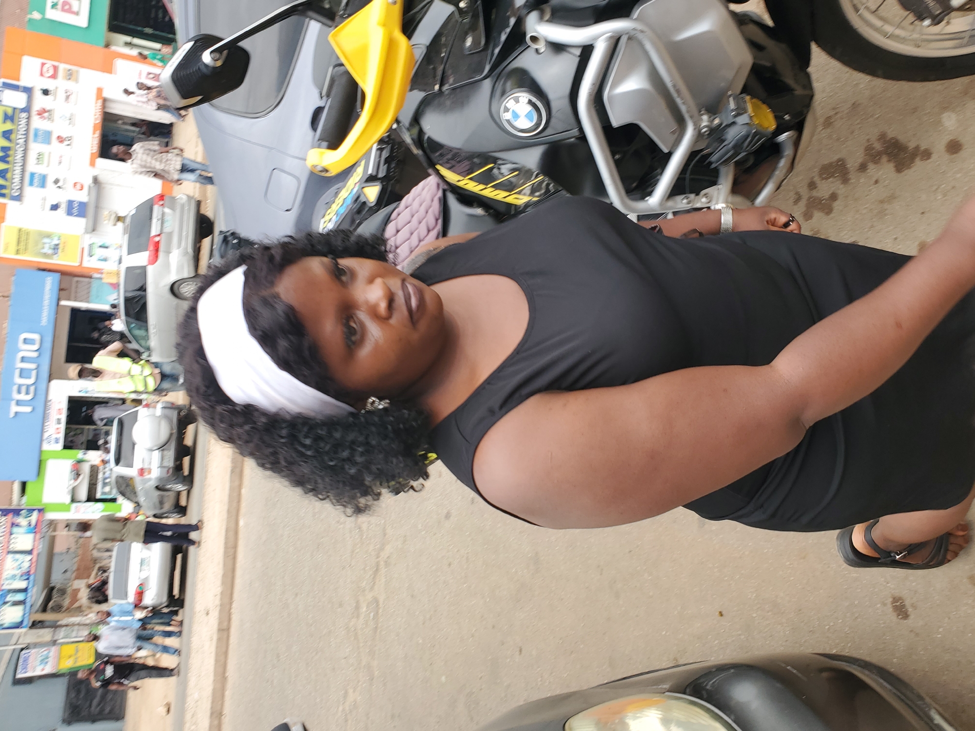 A street scene in Jos, Nigeria, shows a dark-skinned woman in a black dress and white headband standing next to a BMW motorcycle. A vehicle is partially visible at the bottom of the frame. Behind the woman, there are buildings with advertisements and signs including a large "TECNO" sign. Several people are walking on the street. The ground is paved.