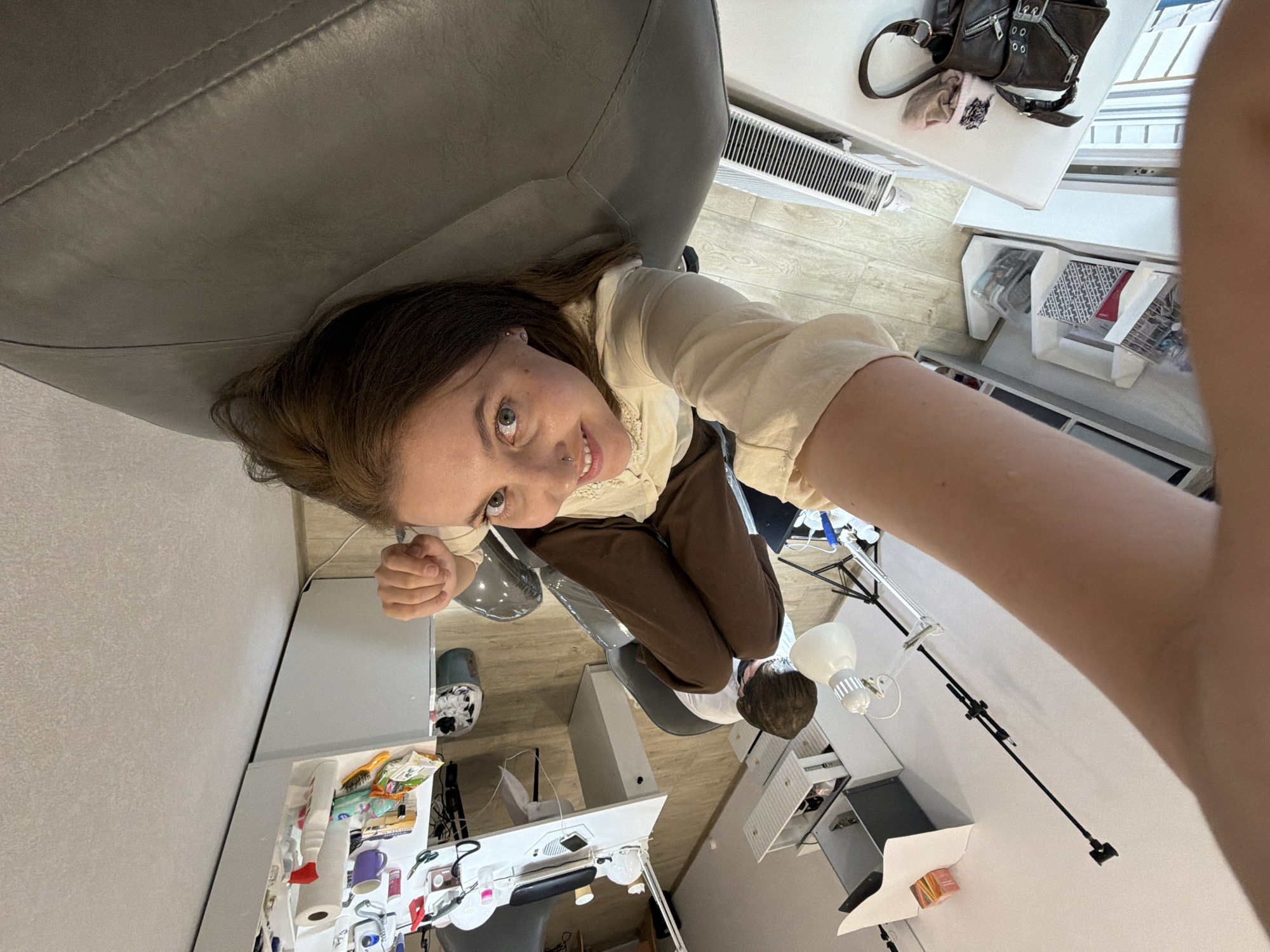 An overhead, wide-angle, selfie-style photograph captures a young woman sitting in a chair, holding the camera with her right hand. Her left hand is raised, touching her forehead. She is wearing a light-colored long-sleeved shirt and brown pants. Her hair is brown and pulled back. She has blue eyes, a nose ring, and a pierced eyebrow. The background appears to be an indoor setting, possibly a home or studio, with a cluttered desk or workspace visible. The room has light-colored walls and a wooden floor. The overall orientation of the image is upside down relative to a standard perspective, suggesting the photo was taken with the camera pointed upwards from a seated position. The image does not provide any specific visual cues to determine its location in Izhevsk, Russia.