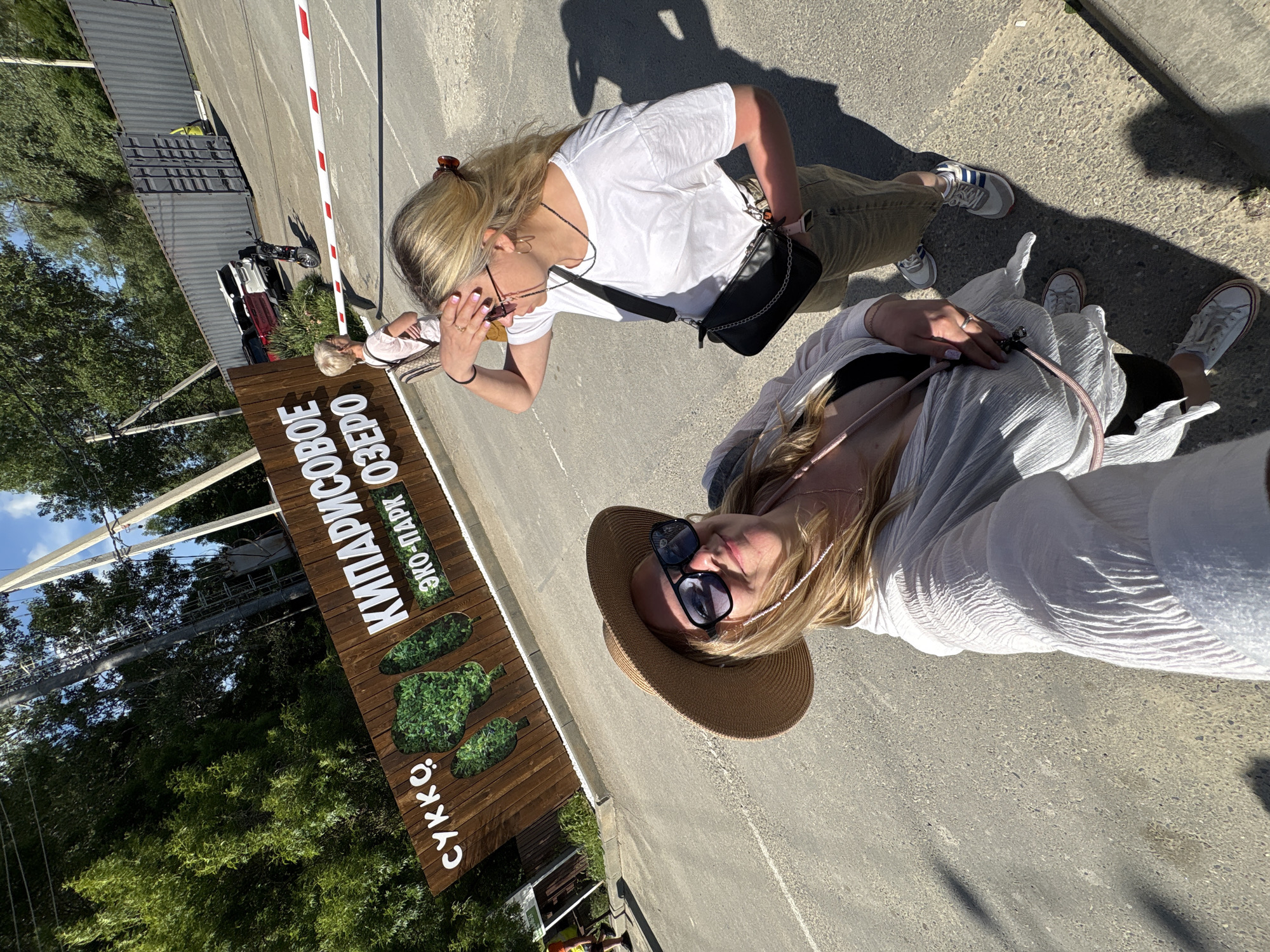 Two women are captured in a daytime outdoor selfie, standing on an asphalt or concrete surface under bright sunshine, indicated by strong shadows. The image is taken at an entrance or checkpoint, marked by a red and white striped barrier arm and a prominent wooden sign.
The woman on the right, facing the camera, is taking the selfie. She has long blonde hair, wears a wide-brimmed straw hat, dark sunglasses perched on her nose, and a white, sheer long-sleeved top over a darker top. A delicate necklace is visible, and she wears a ring on her left hand. Her expression is a slight smile.
The woman on the left, with blonde hair tied back, is looking downwards and slightly to her right. She wears a white t-shirt, khaki-colored pants, and light-colored sneakers with dark stripes. A black crossbody bag hangs from her shoulder, and sunglasses are pushed up onto her head.
In the background, lush green trees are visible under a clear blue sky. Behind the barrier, there are industrial-looking structures, possibly shipping containers, and a scooter or moped. A partial view of another person and a car can also be seen further back.
The large wooden sign, which is a key location cue, features green, stylized shapes resembling cypress trees. Text on the sign, written in Cyrillic, reads: "КИПАРИСОВОЕ ОЗЕРО" (KIPARISOVOE OZERO), which translates to "Cypress Lake," and "ЭКО-ПАРК" (EKO-PARK), meaning "Eco-Park." At the bottom, the word "СУККÖ" (SUKKO) is displayed. This indicates the women are at the Cypress Lake Eco-Park in Sukko.