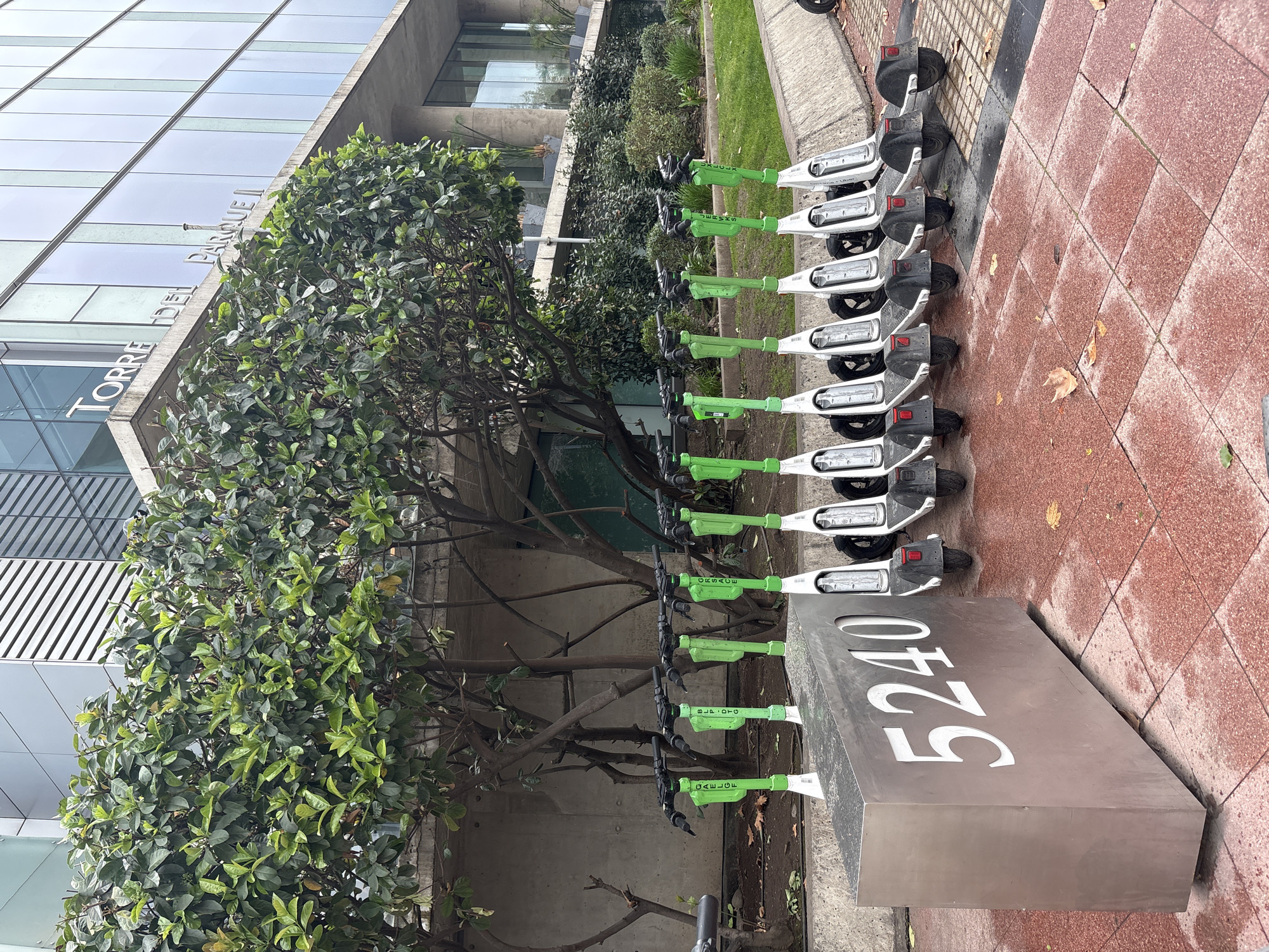 The image depicts a row of electric scooters parked in front of a modern building, identified as "TORRE DEL PARQUE II" in Santiago, Chile. The building features a glass facade and concrete elements, with lush green trees and bushes in the foreground. A silver metallic structure with the number "5240" is positioned to the left of the scooters. The scooters are predominantly white with green accents and handlebars. They are lined up neatly on a reddish-brown tiled walkway. The scene appears to be an outdoor urban environment, possibly a business district or a public plaza.