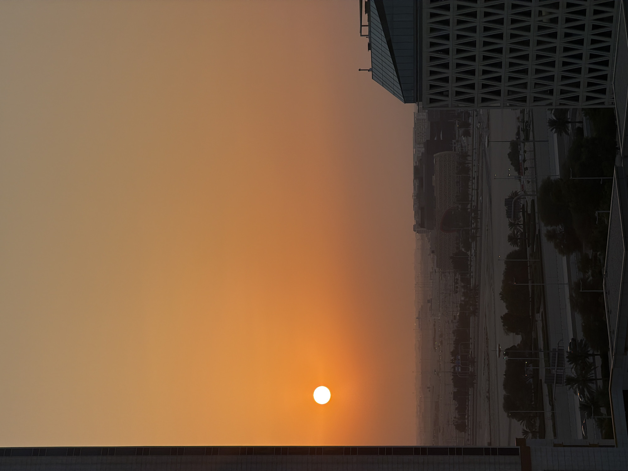 The image depicts a sunset scene in Lusail, Qatar, characterized by a clear sky displaying a gradient of orange hues. The sun, a bright white orb, is visible in the left half of the frame, casting a warm glow. In the foreground, a modern building with a distinctive white patterned facade is partially visible on the right. To the left, a tall, dark skyscraper stands prominently. The middle ground reveals a cityscape with various buildings, roads, streetlights, and scattered trees, suggesting an urban environment. The overall atmosphere is serene, with the setting sun creating a dramatic and peaceful ambiance over the city.