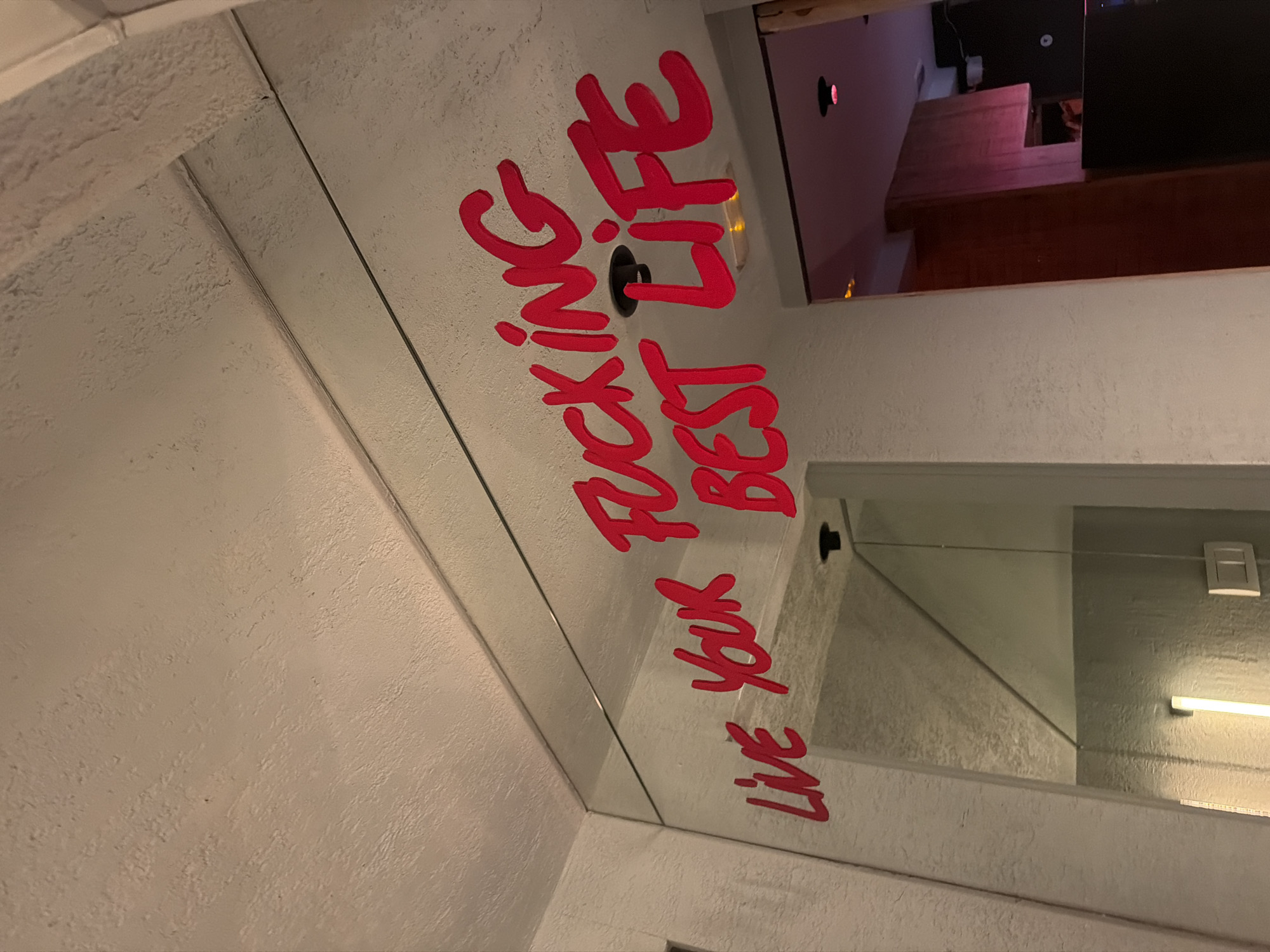 The image shows a bathroom mirror in what appears to be a commercial establishment. Bold, red graffiti-style lettering is applied directly on the mirror’s surface, reading "LIVE YOUR FUCKING BEST LIFE”. The reflection in the mirror provides views of the interior of an adjacent space, where furniture, lighting, and architectural details are visible. Based on similar images, the location is likely in Barcelona, Spain.